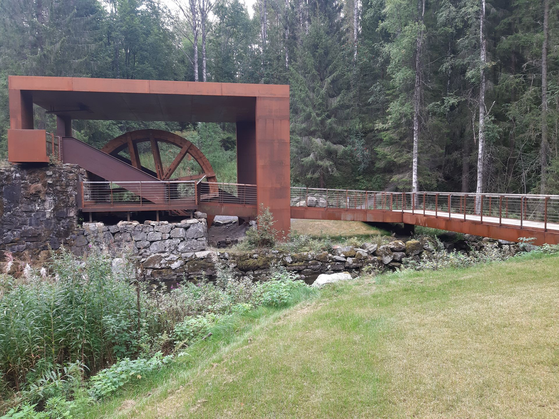 Rusten metallkonstruksjon med vannhjul og bro i skogsområde.
