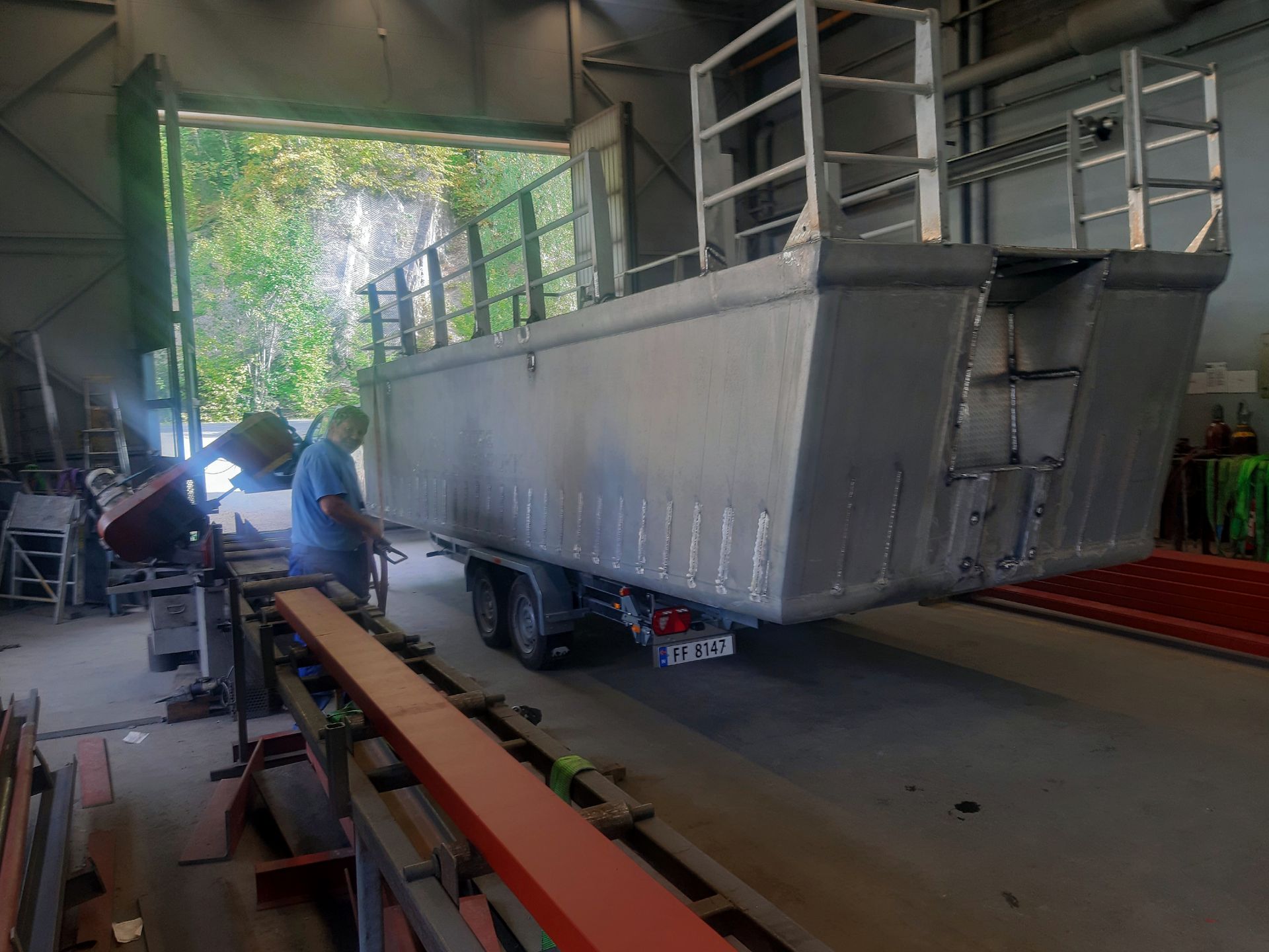 En arbeider sveiser på en stor aluminiumsbåtramme på en tilhenger inne i et verksted.