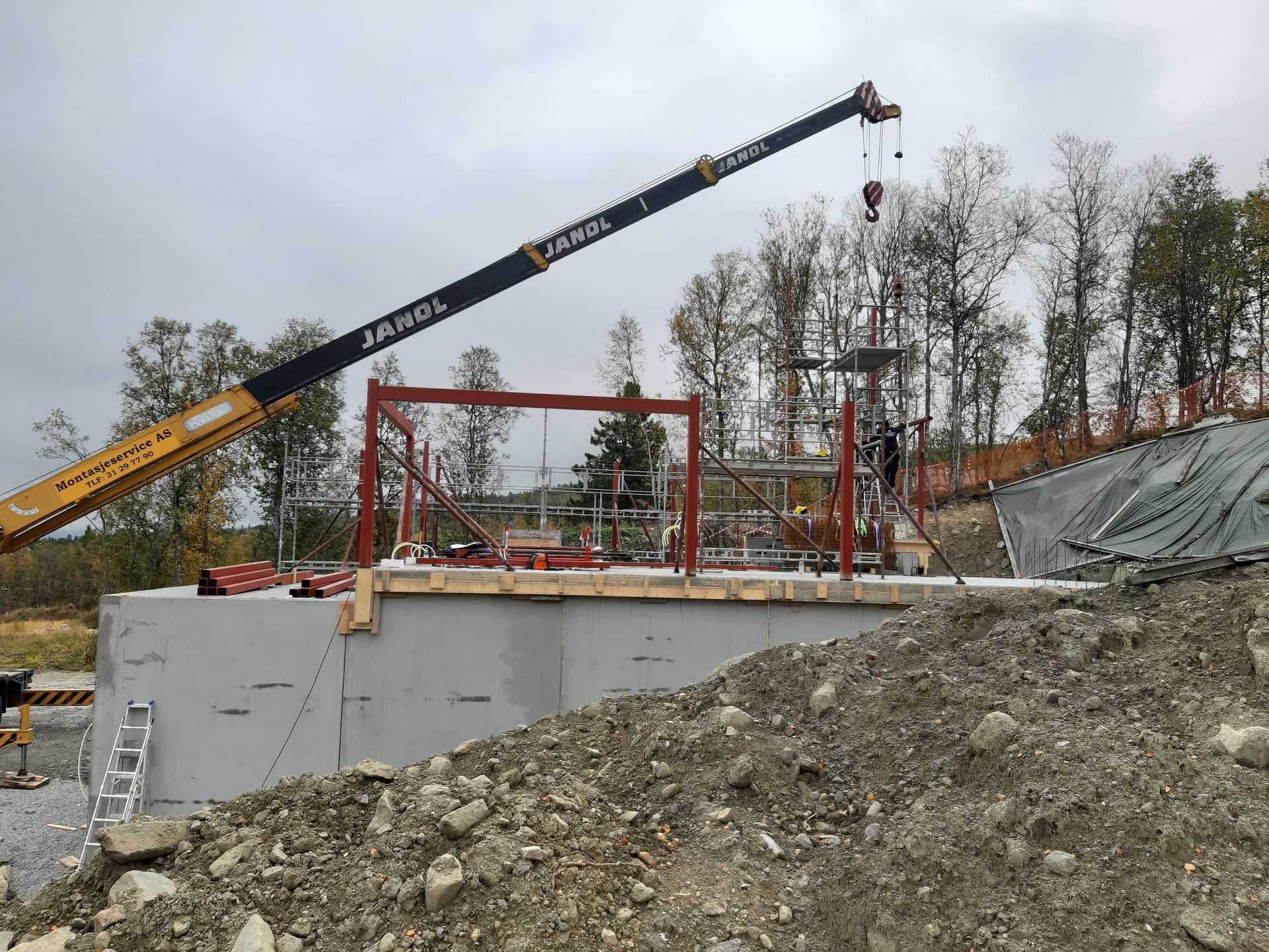Kran løfter stålramme opp på en betongkonstruksjon på en byggeplass; overskyet dag.