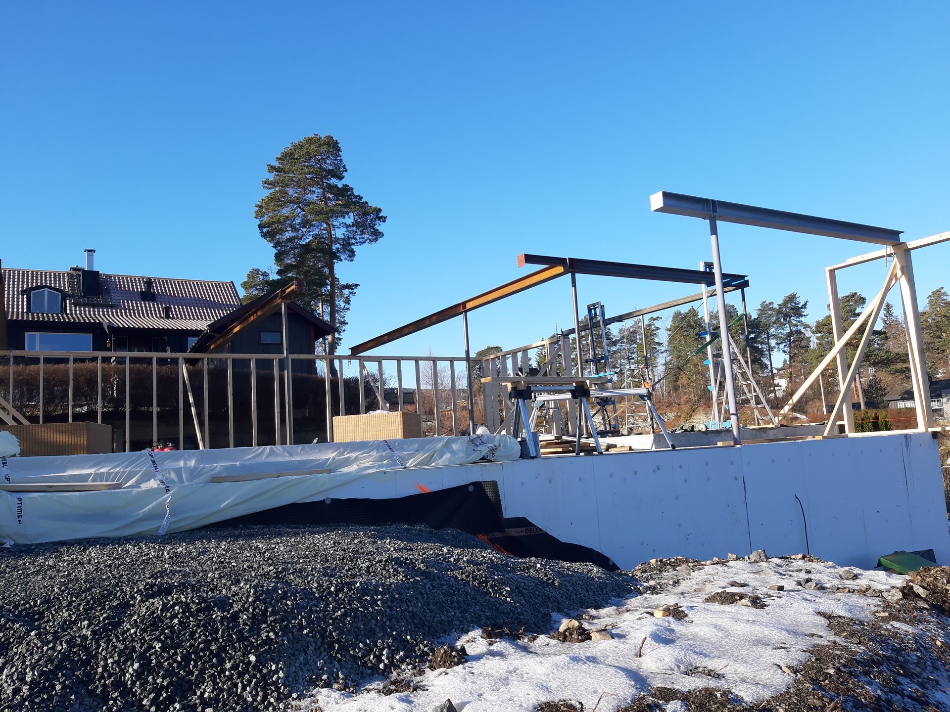 Byggeplass med synlig trekonstruksjon, hvite paneler og grus, med et hus i bakgrunnen under en blå himmel.
