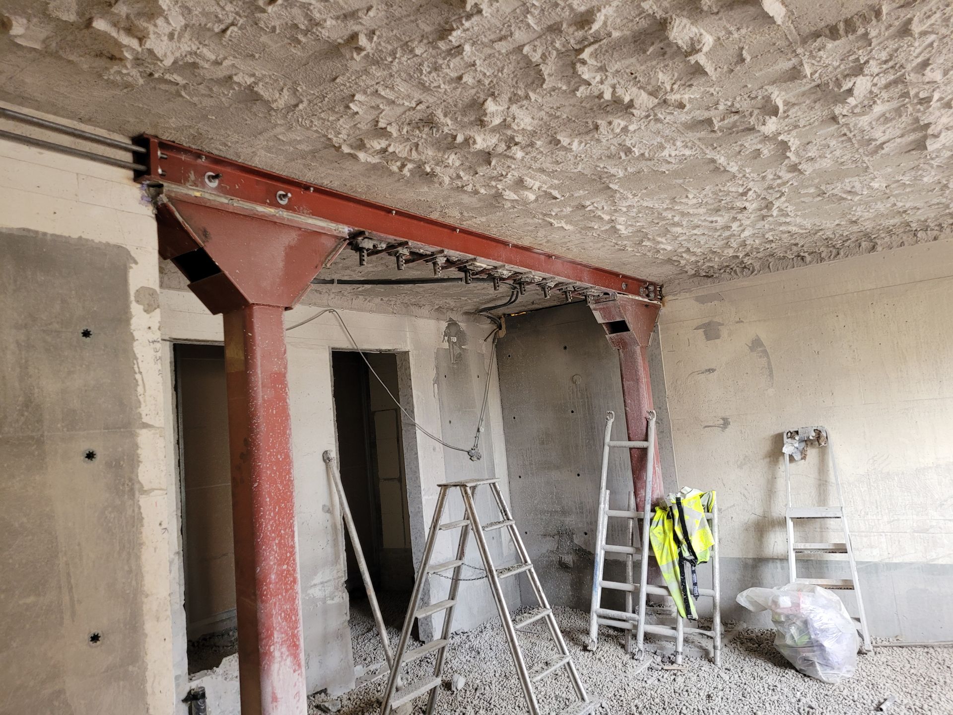 Byggeplass: Rød stålbjelke støtter taket i et rom med synlige vegger og rusk. Stiger til stede.