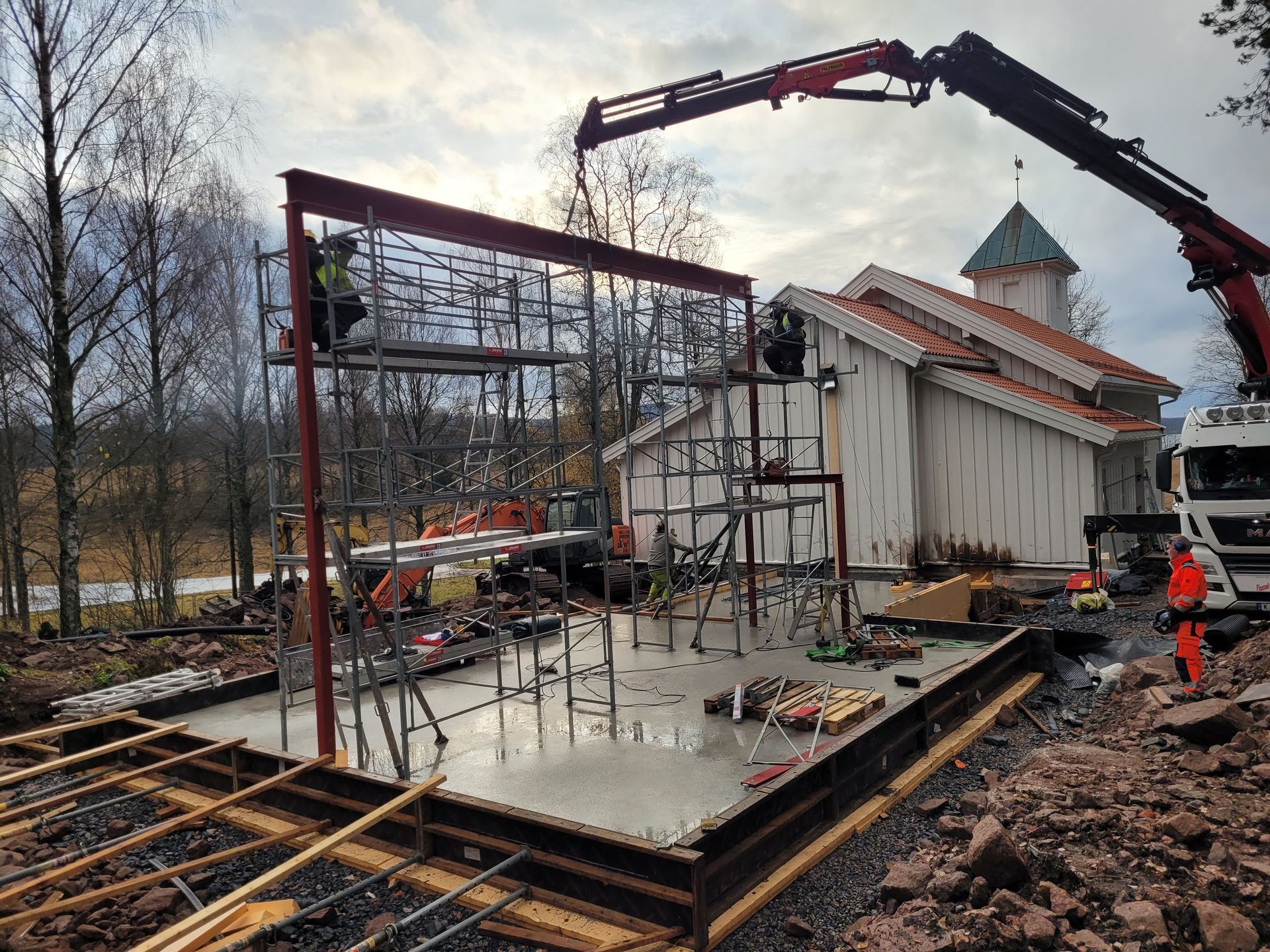 Byggeplass med kran som løfter stålramme, ved siden av en hvit trebygning med spir. Arbeidere til stede.