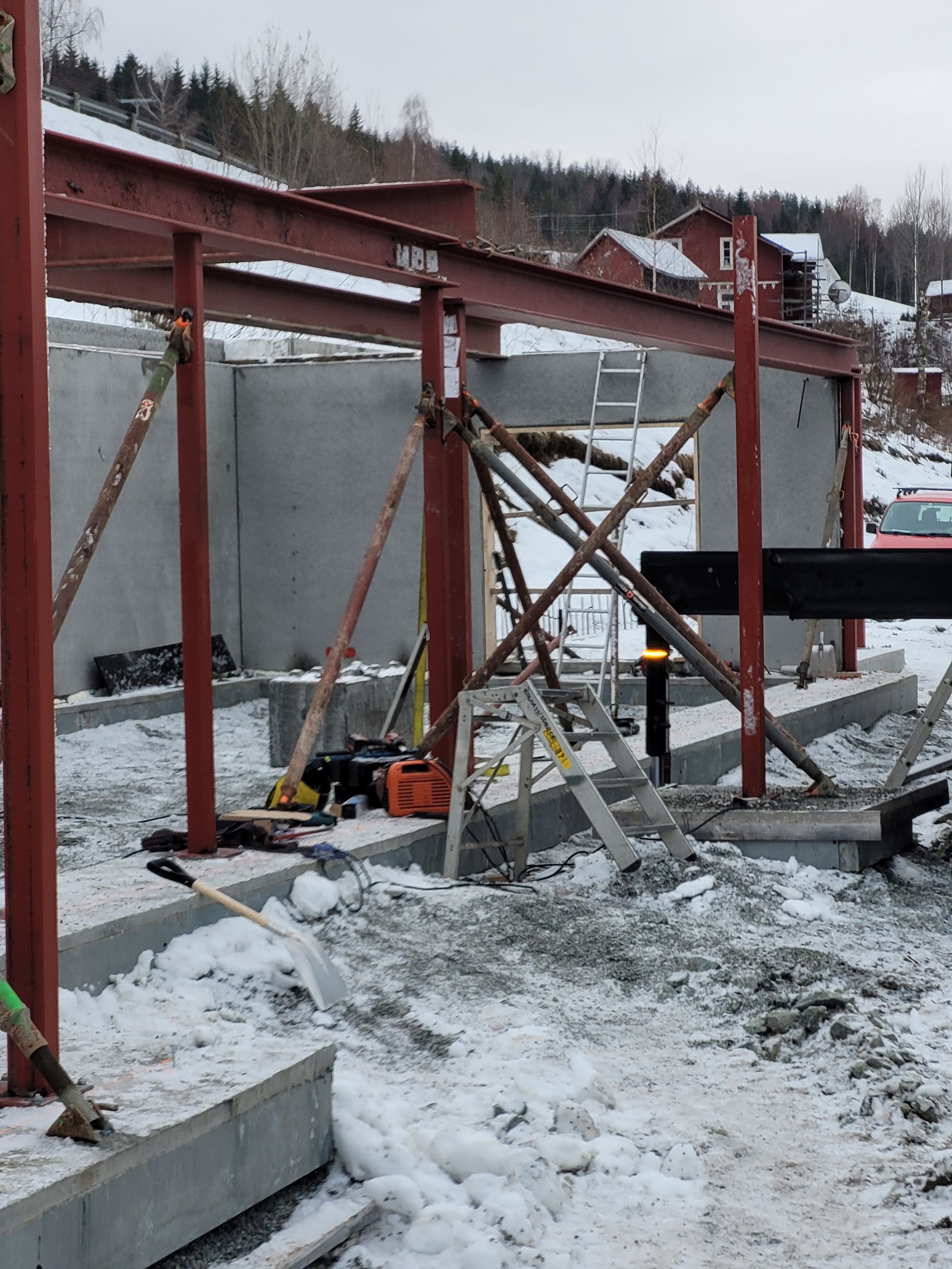Byggeplass med røde stålbjelker, betongvegger og snø. En stige lener seg mot konstruksjonen.