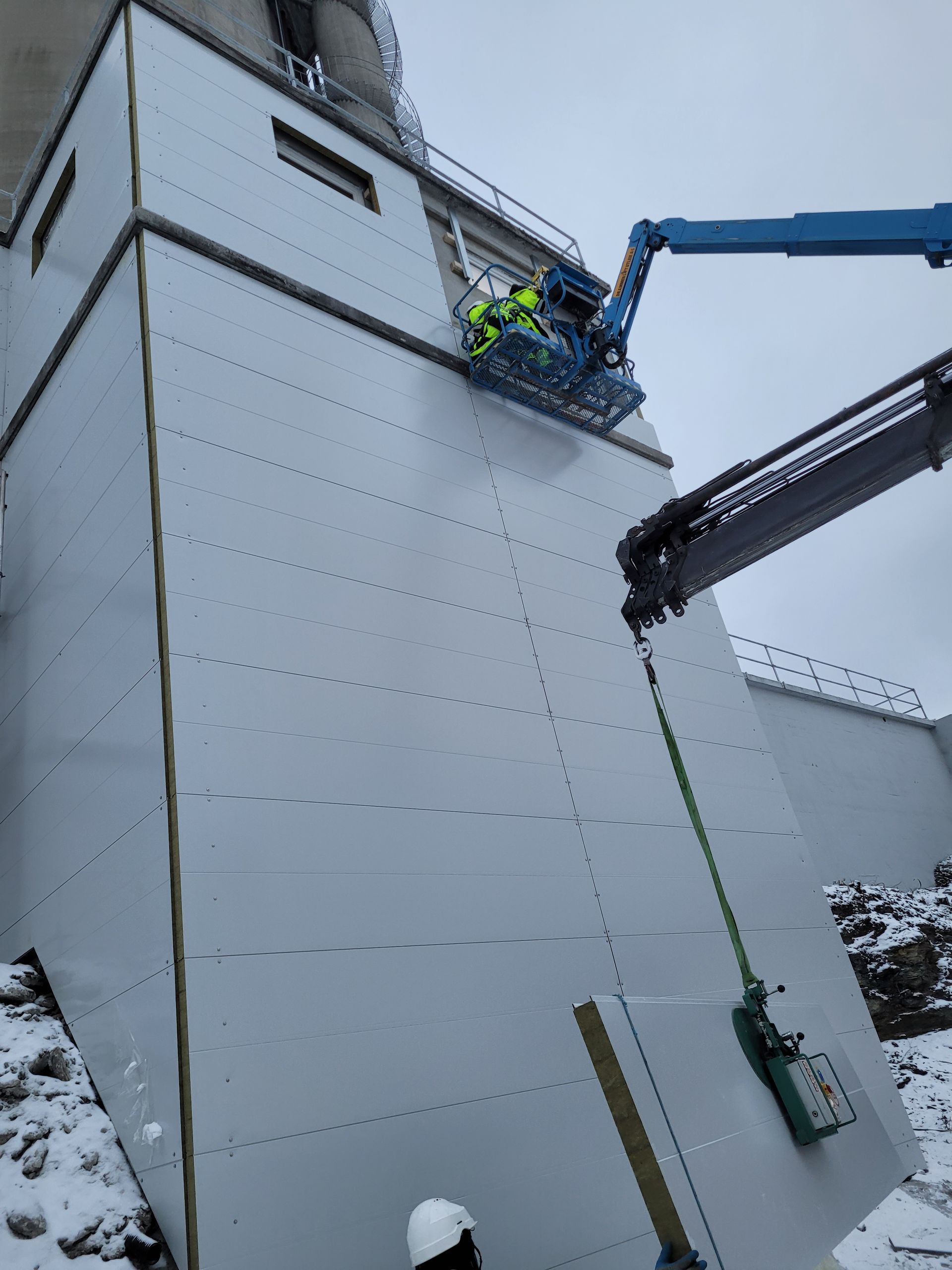 Bygningsarbeidere på en heis fester paneler til en stor hvit bygning på snødekt underlag.