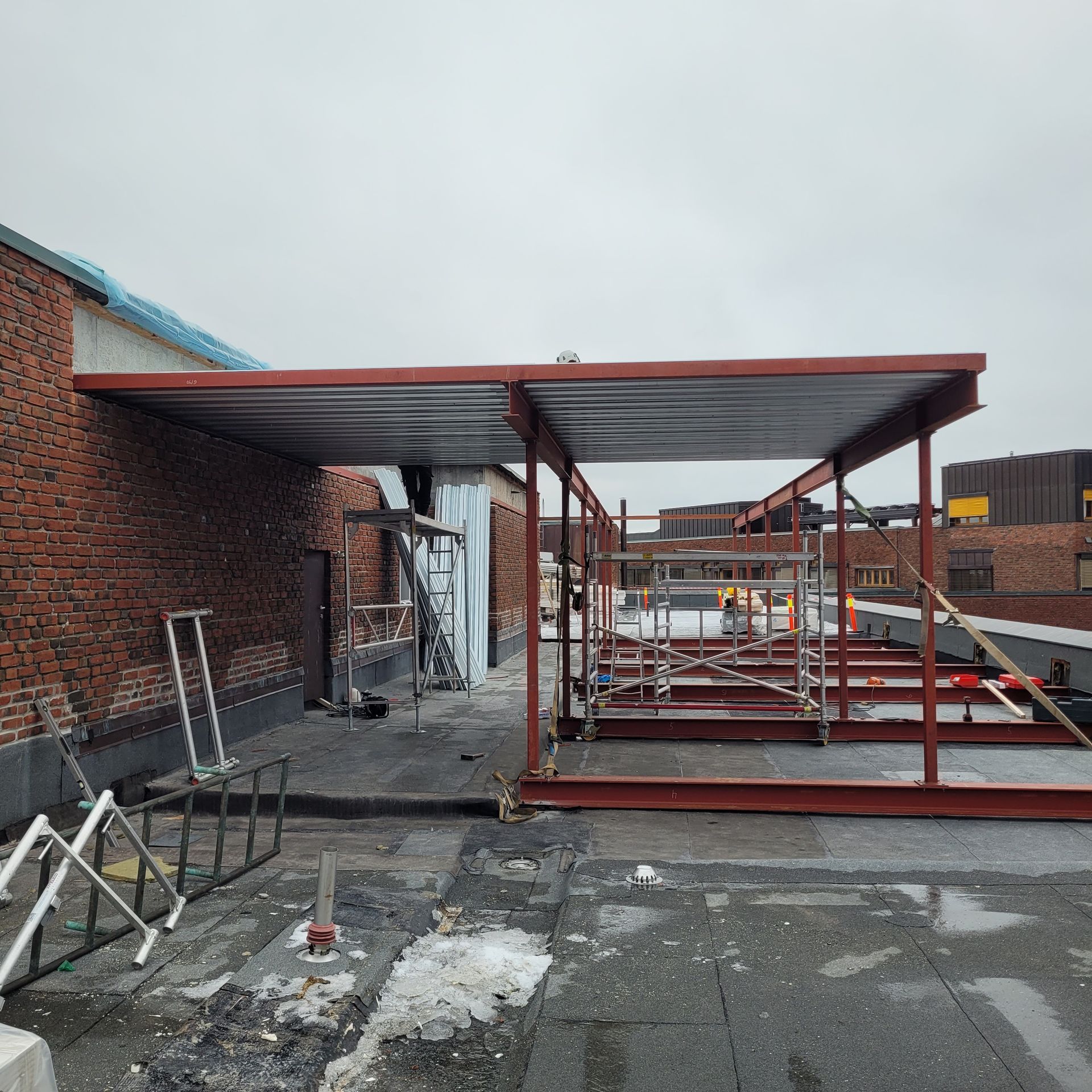Takkonstruksjon med rødt stålrammeverk og en murbygning på en overskyet dag.