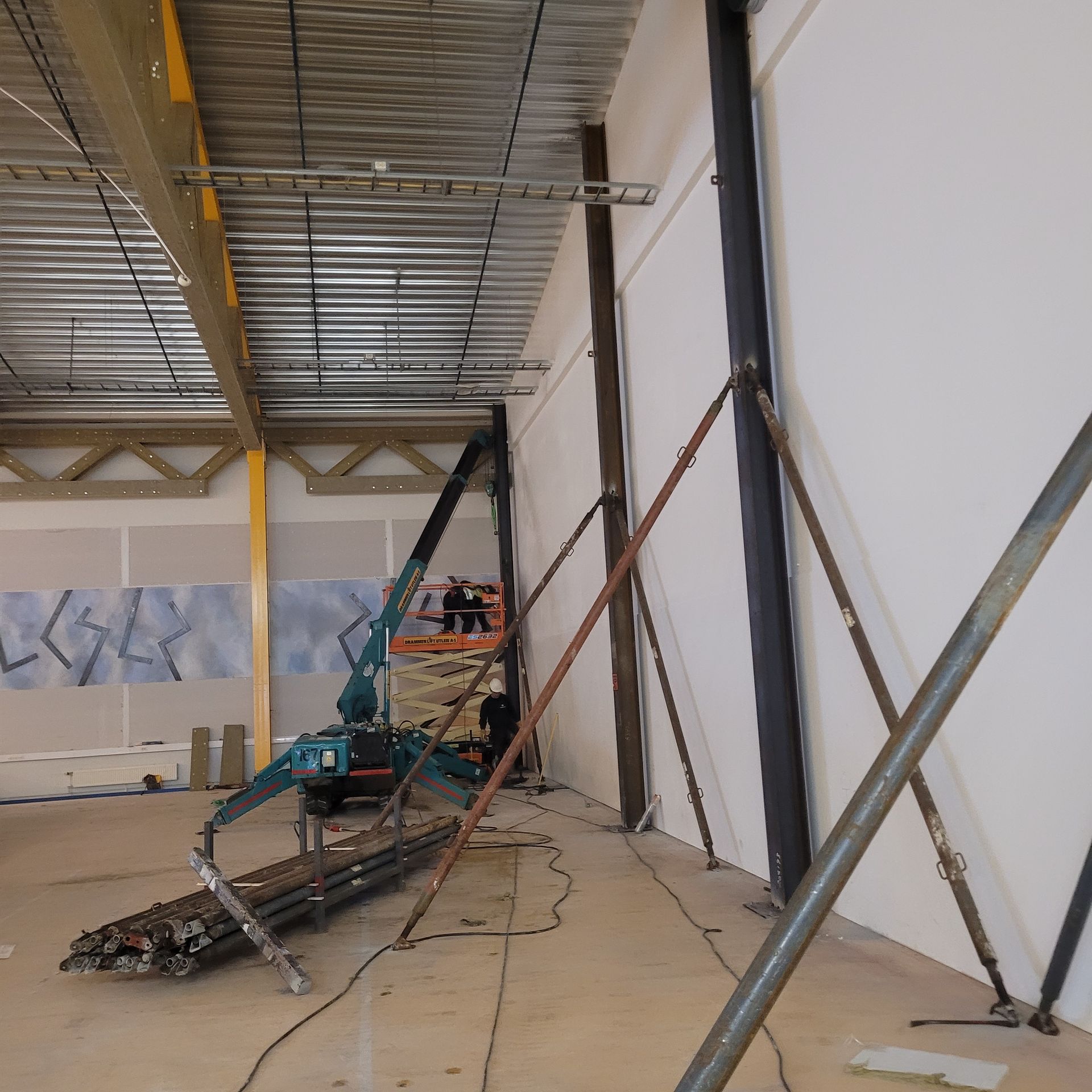 Interiør på byggeplass: arbeidere på en heis, stålbjelker, midlertidige støtter, hvit vegg, metalltak.