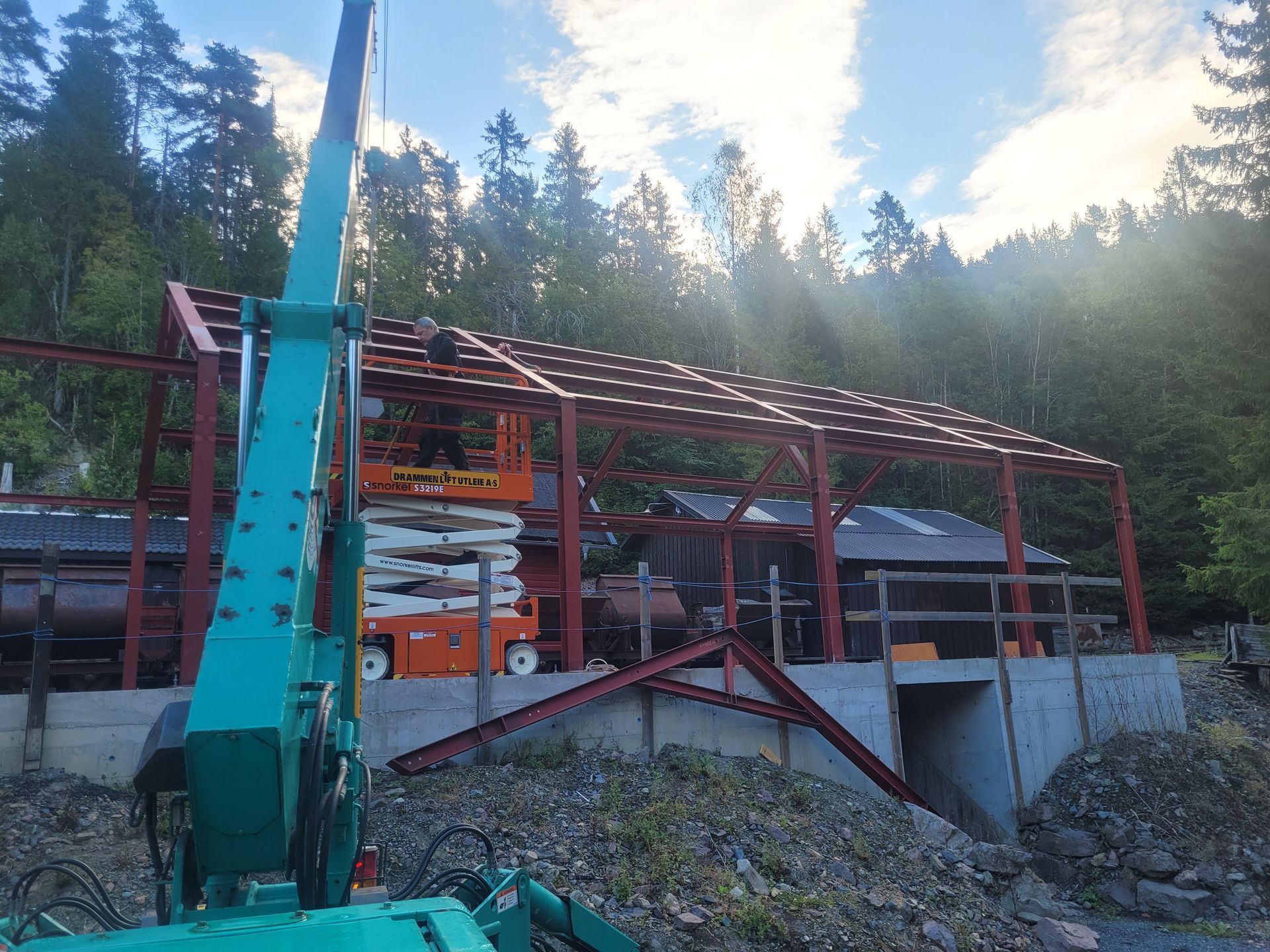 Byggeplass: Rødt metallrammeverk for et skurtak. En løfteplattform og maskineri er til stede, med trær i bakgrunnen.