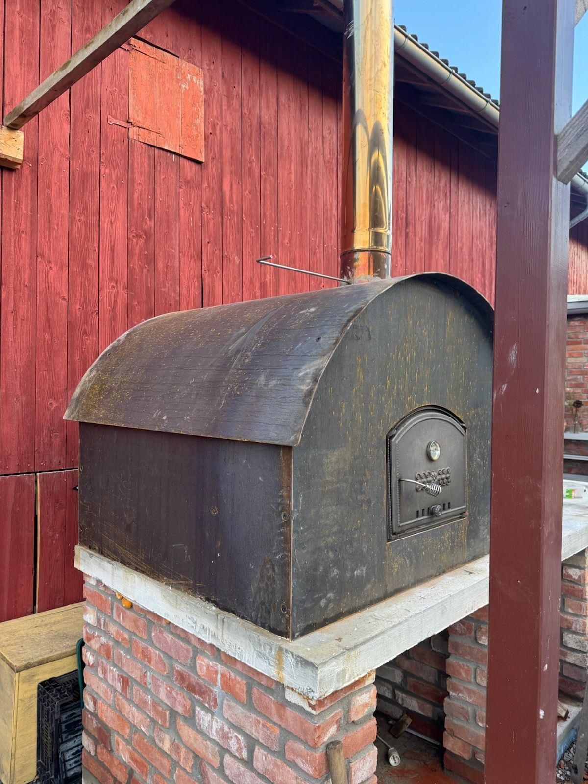 Pizzaovn med metallhette og skorstein, mot rød trevegg.