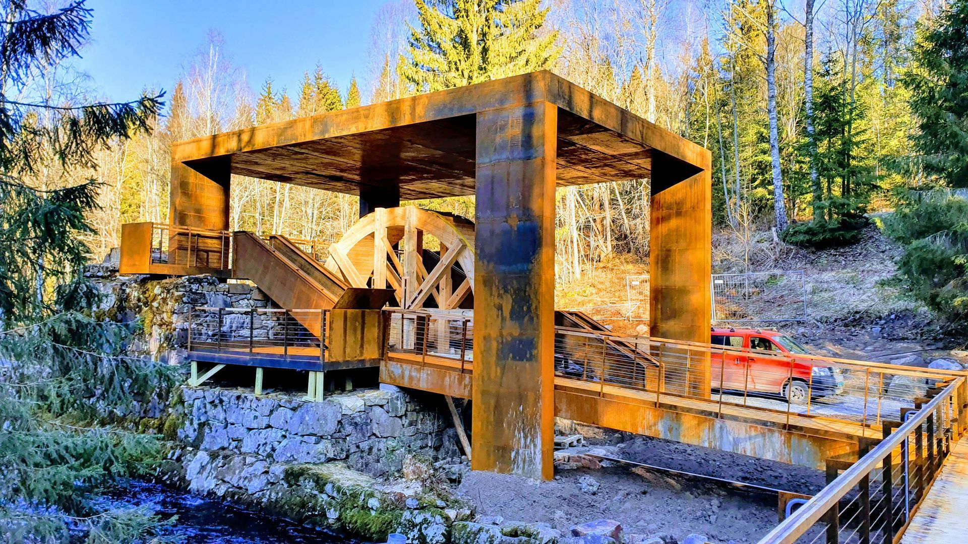 Rusten metallkonstruksjon med vannhjul og tregangvei, plassert i en skog.