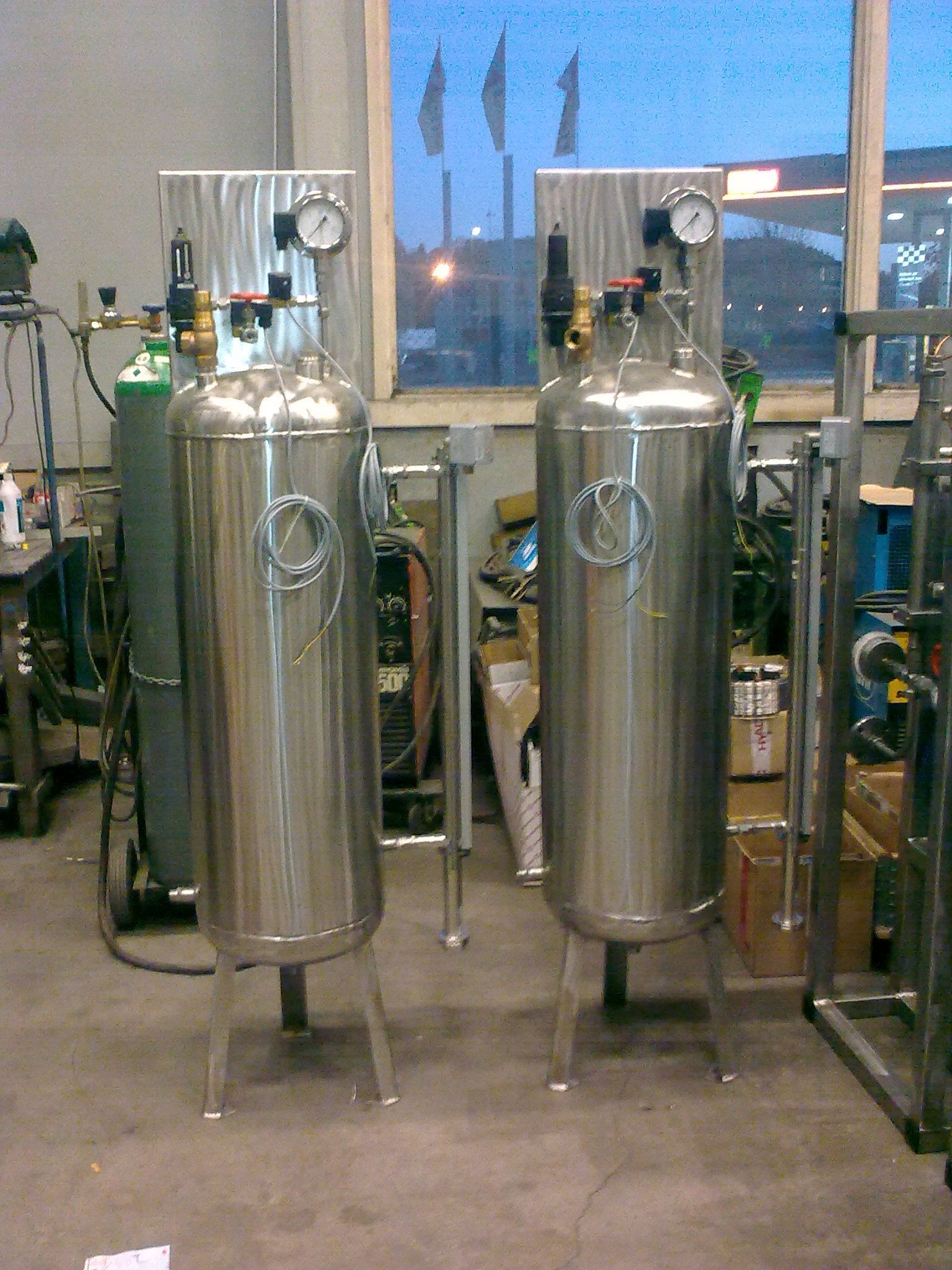 To høye, skinnende, sylindriske metalltanker med målere og regulatorer, innendørs, industrielt miljø.