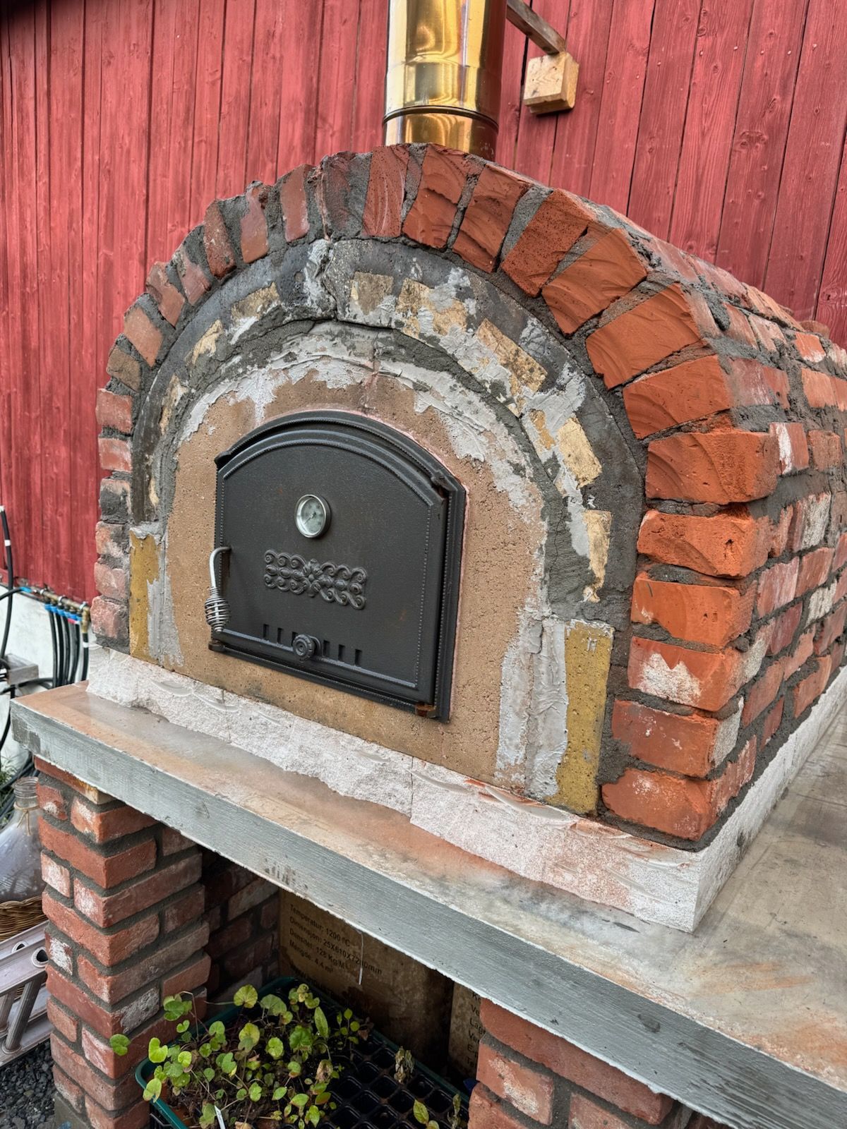 Mursteinspizzaovn med metalldør og skorstein mot en rød vegg.