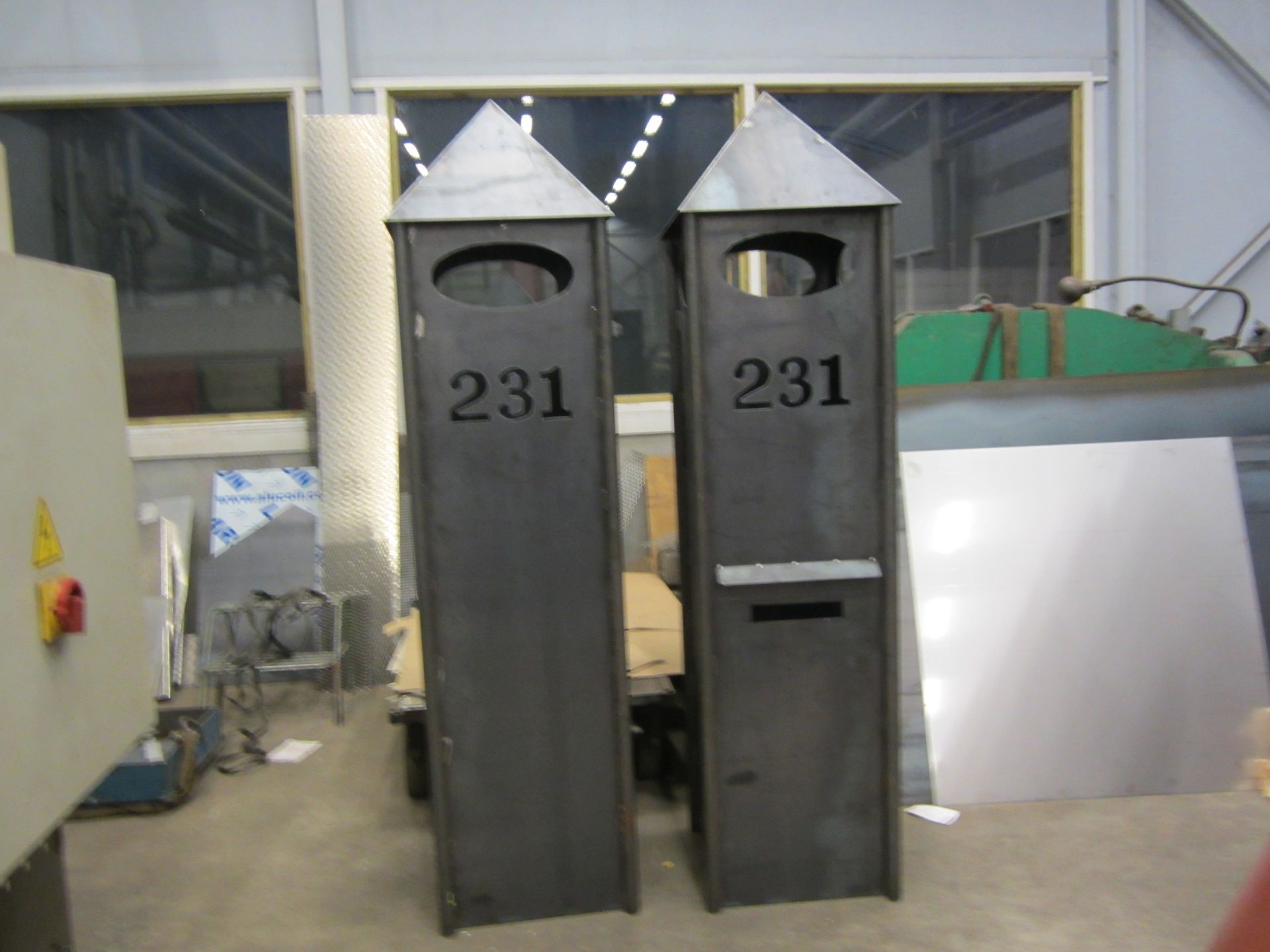 To høye, mørke metallpostkasser med spisse tak, nummer 231, og ovale åpninger. Inne i et verksted.