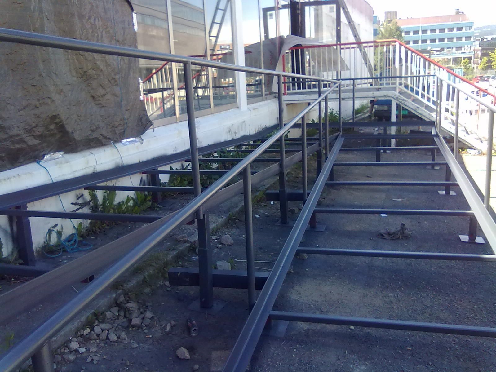 Rampe og trapp med metallrekkverk, som fører opp til en bygningsinngang. Grått og svart.