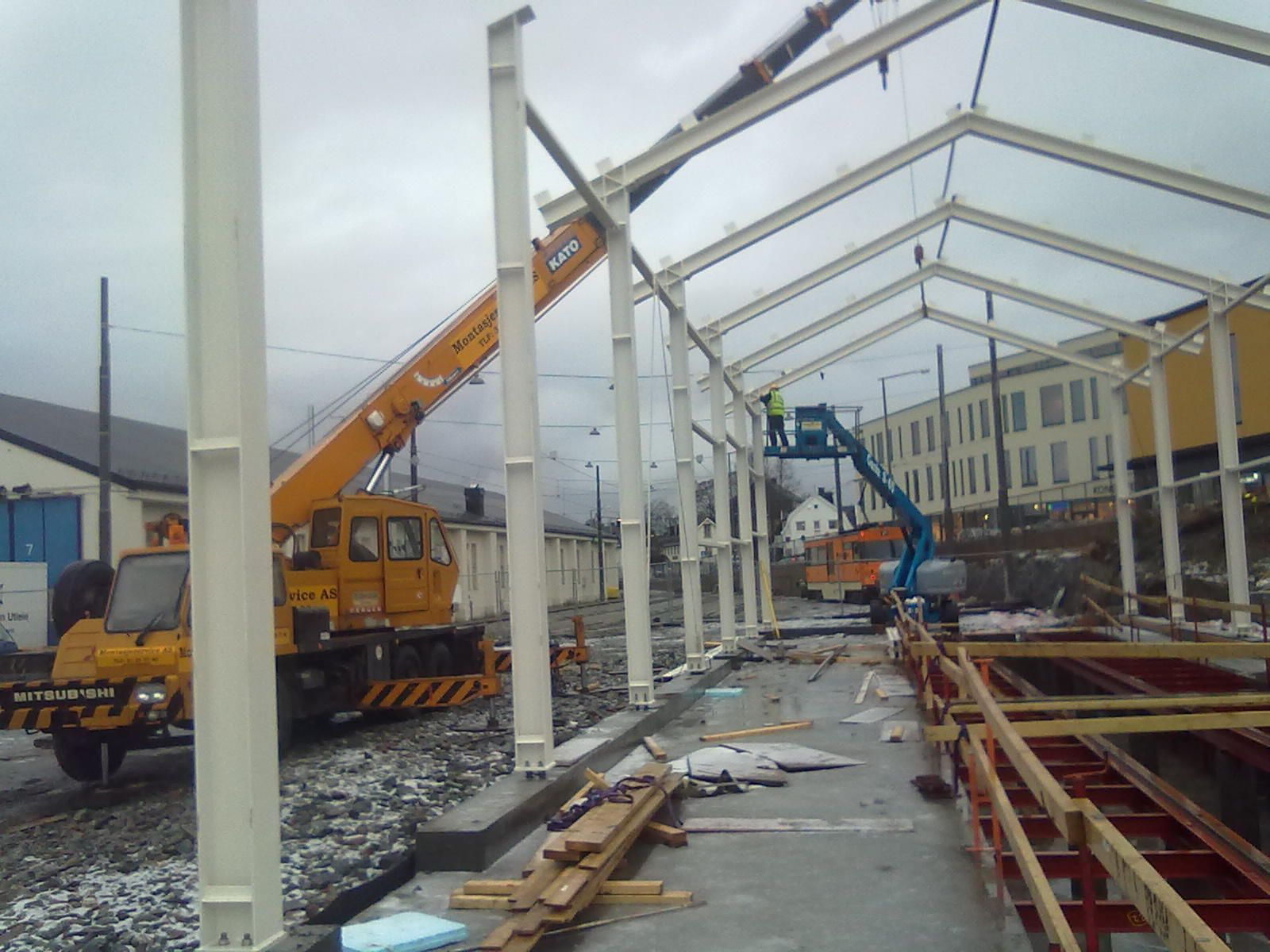 Byggeplass med kran som løfter stålbjelker; en arbeider på en heis. Grå himmel, gult og blått maskineri.