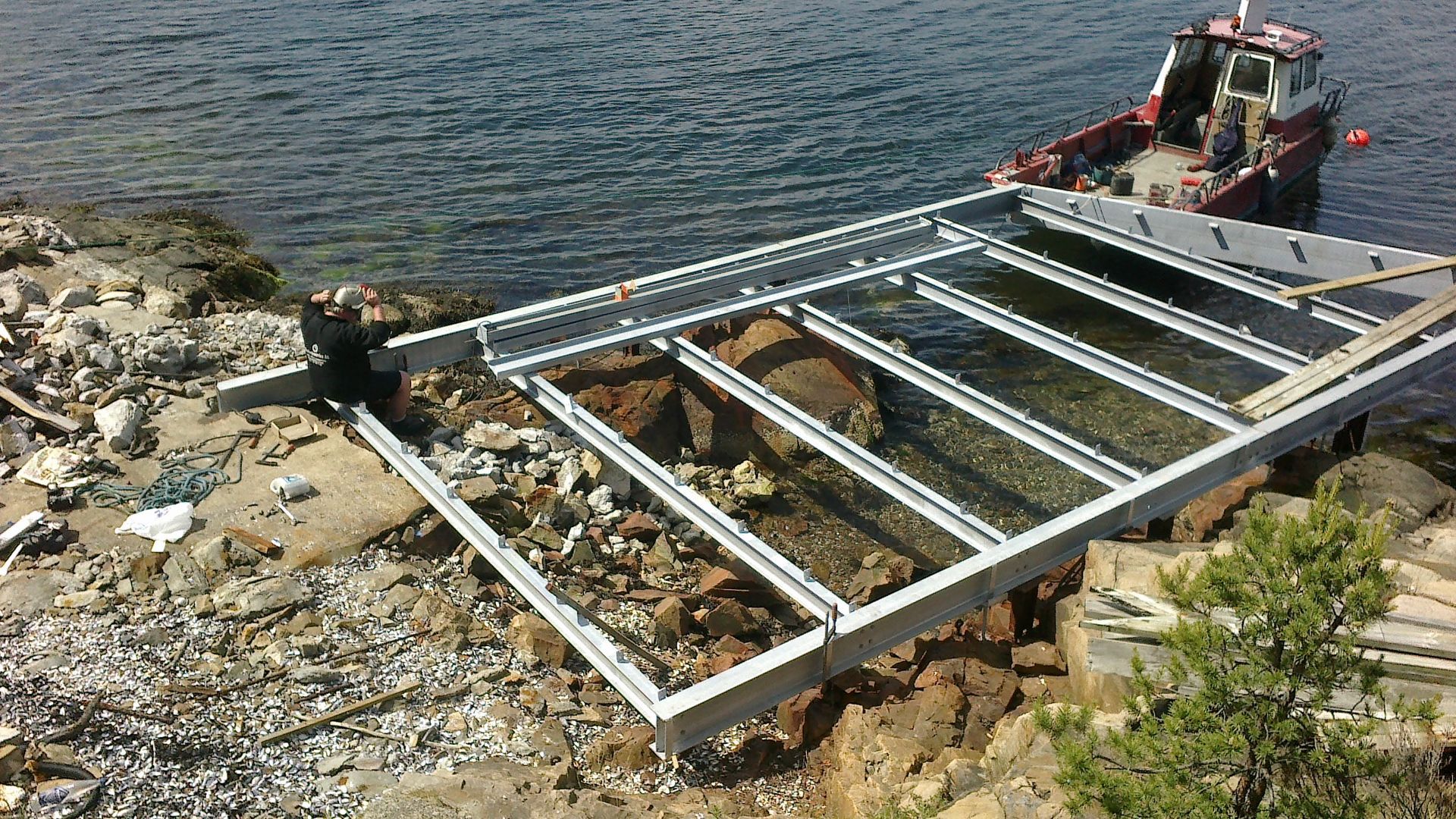 Metallkonstruksjon bygges på en steinete strand nær vann, med en båt i bakgrunnen.