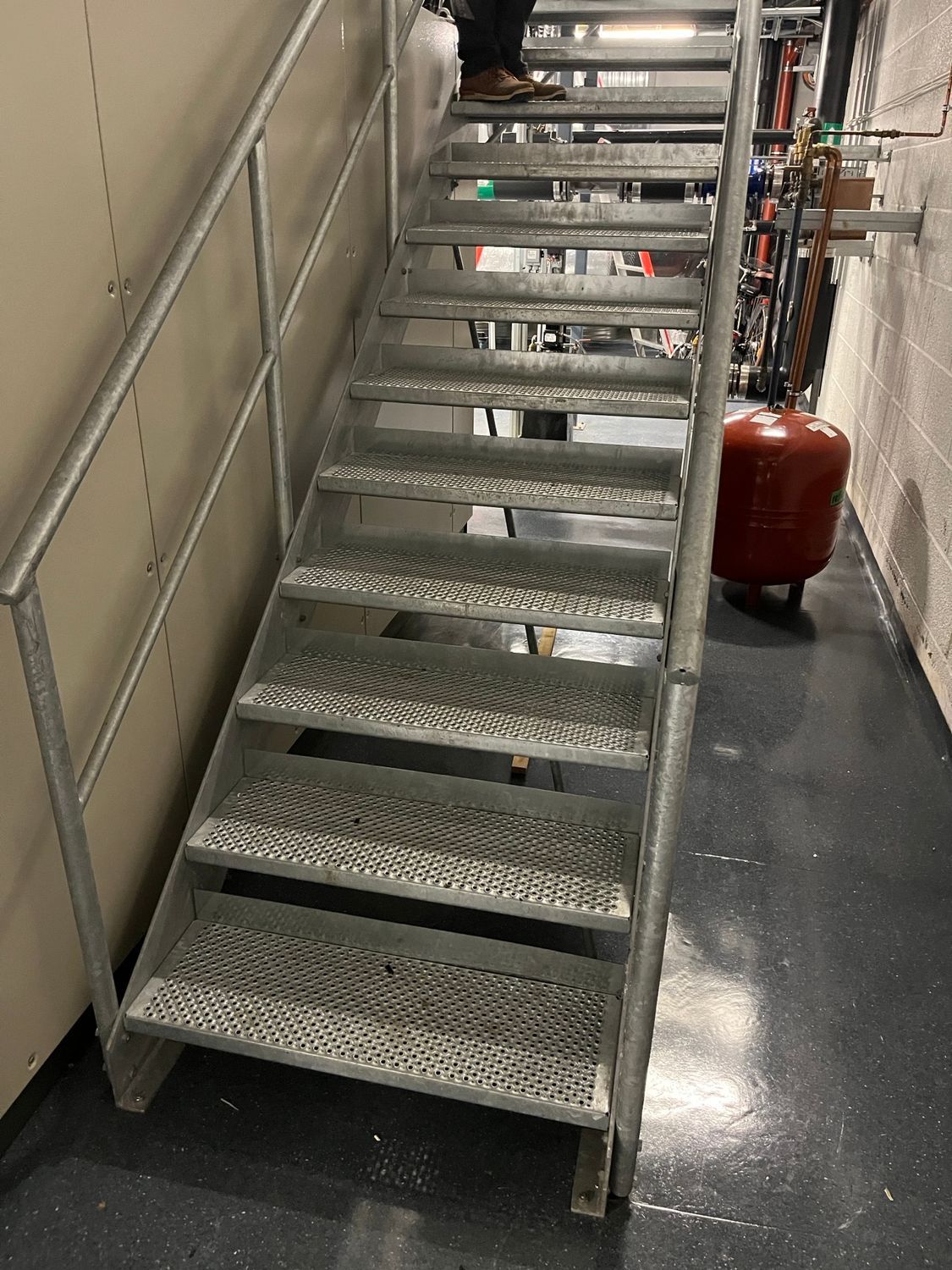 Metalltrapp fører oppover, person går oppover. Mørkt gulv, utstyr og en rød tank er synlige.