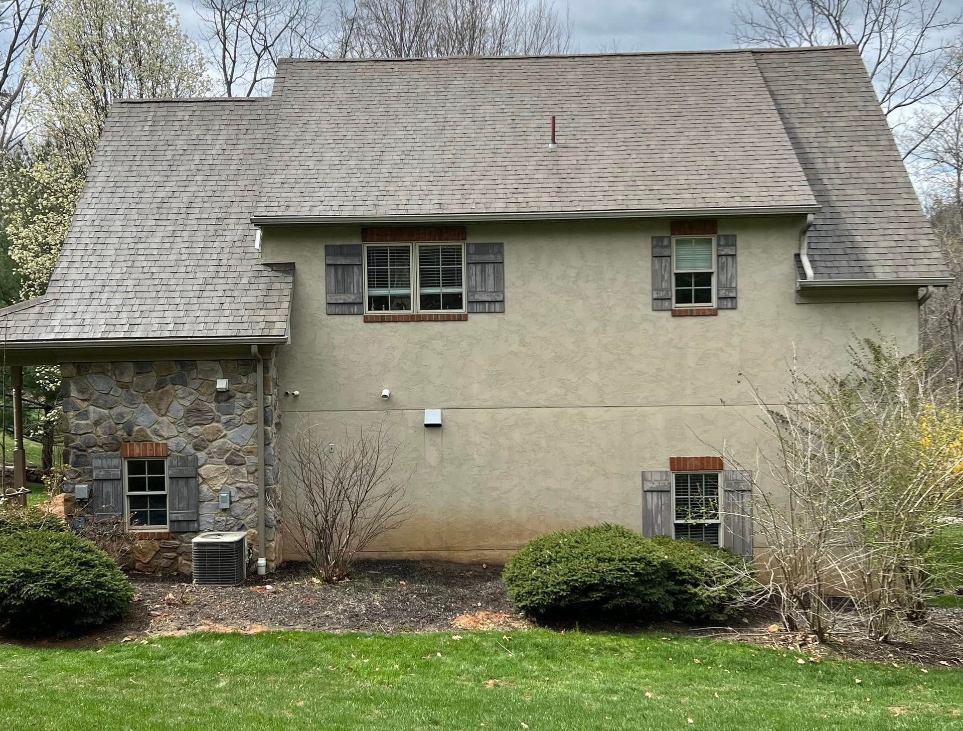 Two-story house with a stone facade, beige stucco, wooden shutters, and a weathered shingle roof.