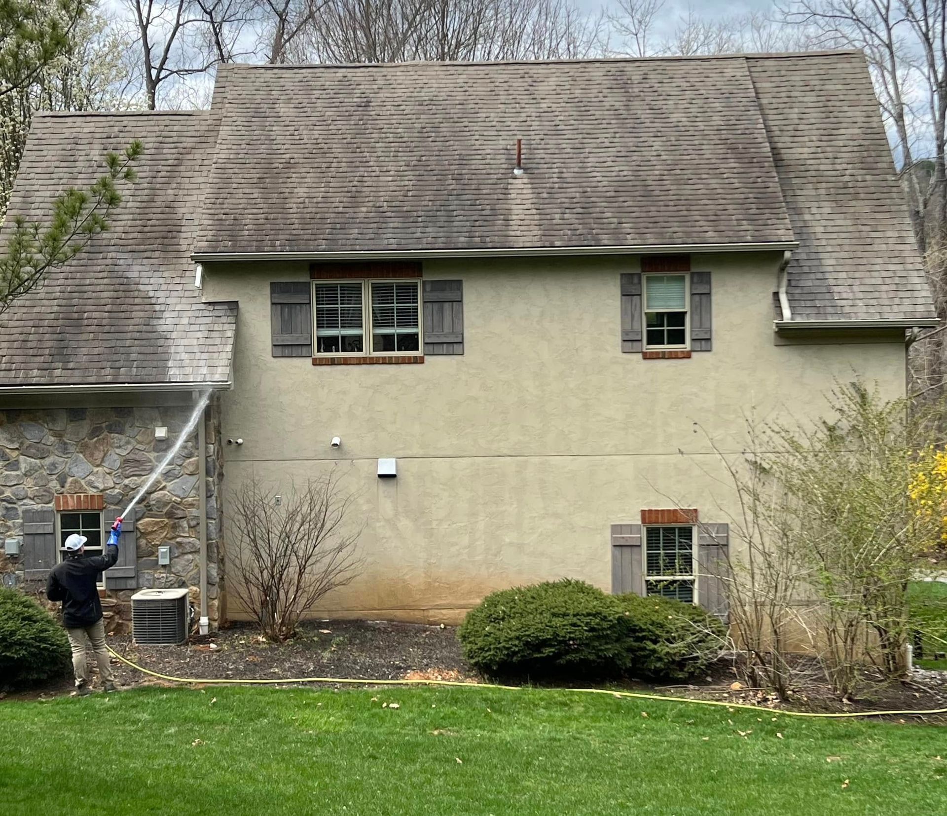 Person pressure washing a house roof; tan exterior, brown roof, green grass.