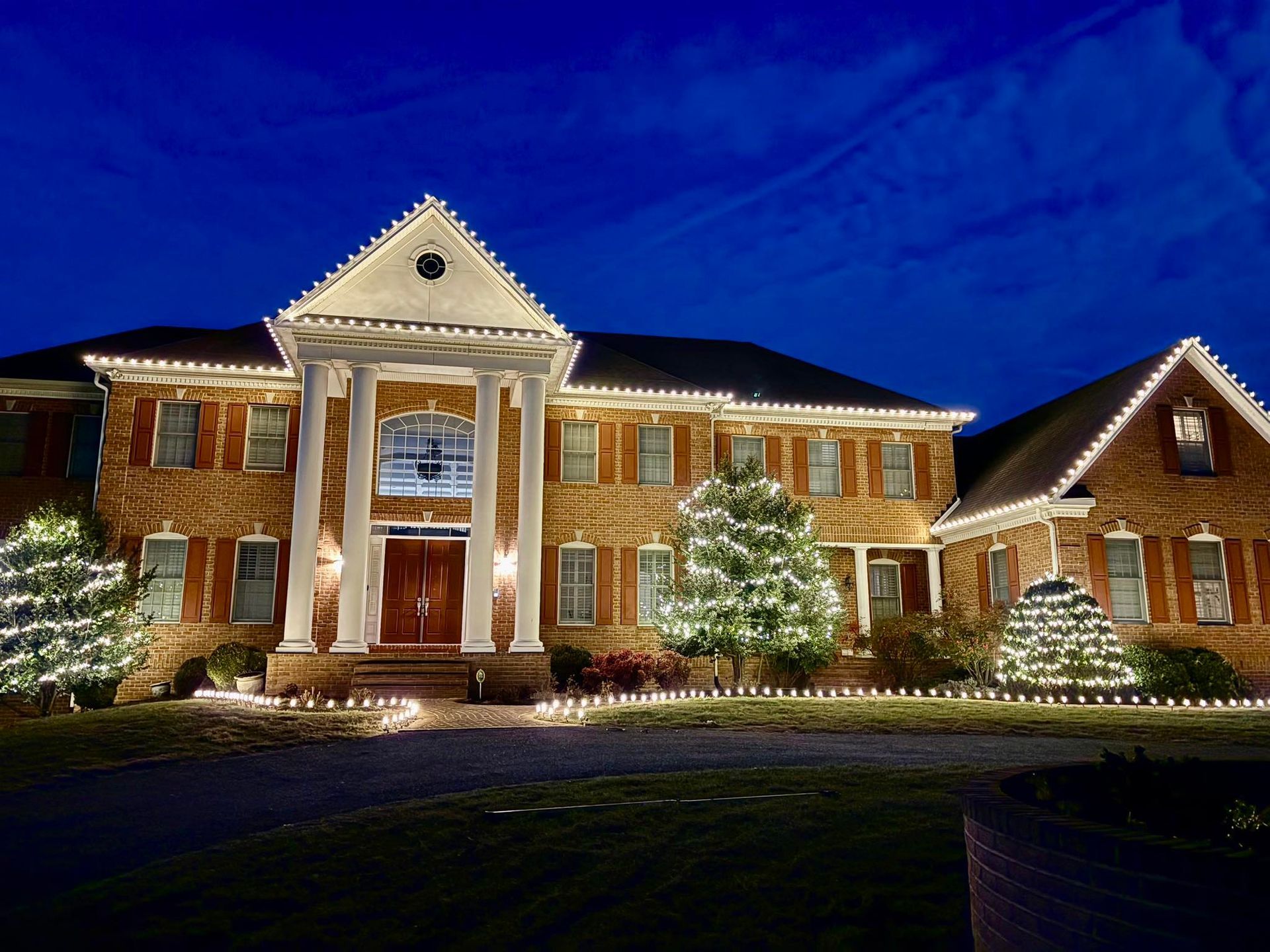 Large brick home with Christmas lights outlining roof, windows, and bushes against a dark blue sky.