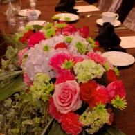 Bright mixed floral centerpiece with pink roses on a dining table, with cups and plates in the background