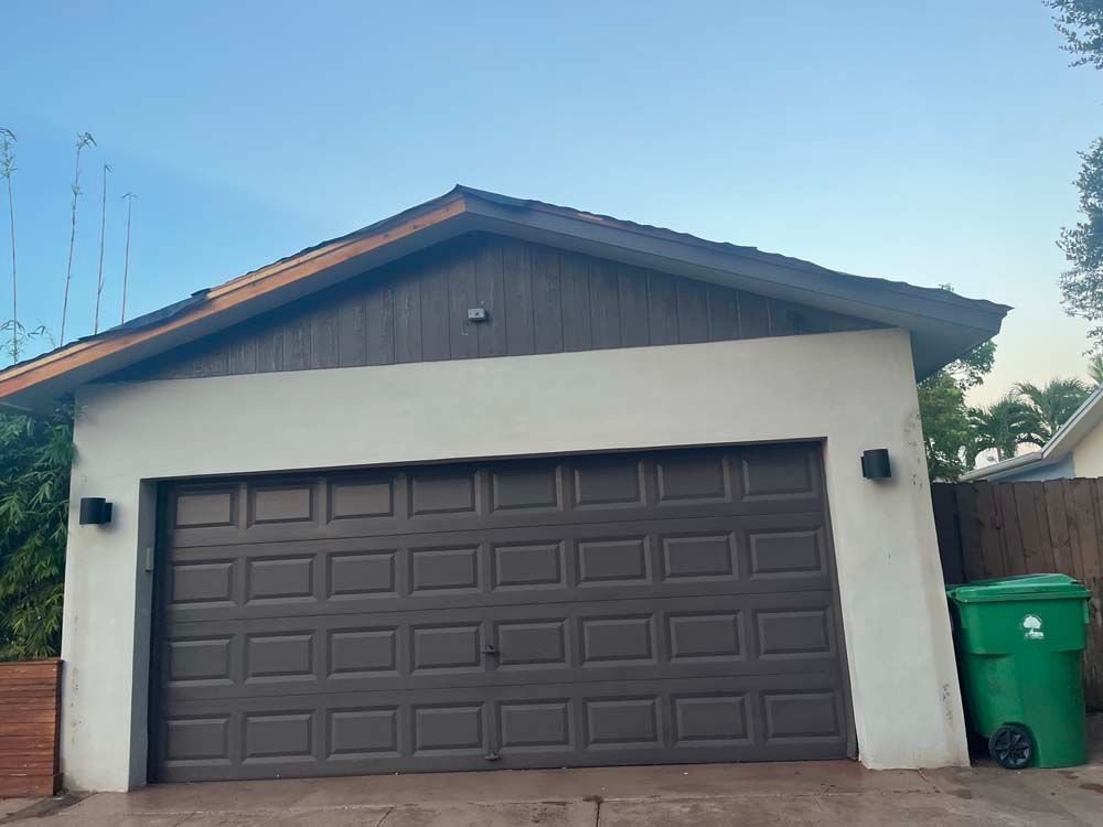 A garage with a brown door and a green trash can in front of it.