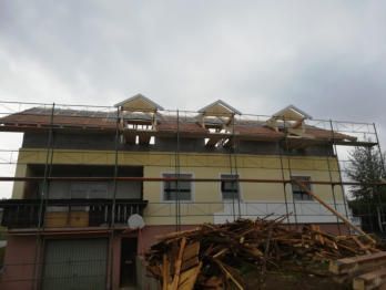 Haus im Bau mit Gerüst, Dachgauben und Holzstapel. Gelbe und rosa Fassade.