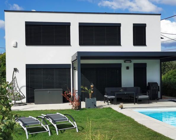 Modernes weißes Haus mit dunklen Jalousien, Terrasse, Pool und Liegestühlen auf dem Rasen.