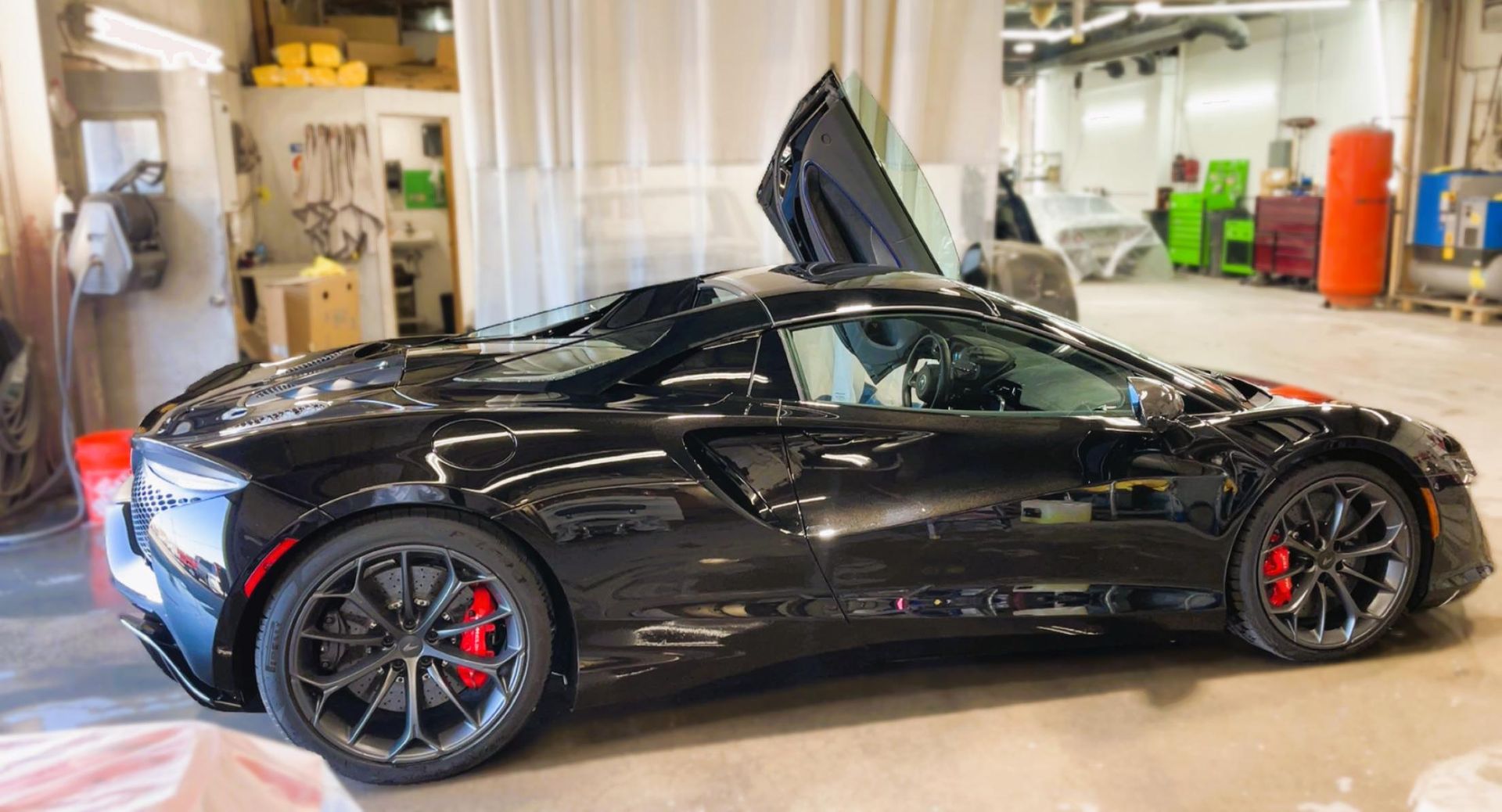 Black sports car with butterfly doors open in a workshop. Red brake calipers visible.