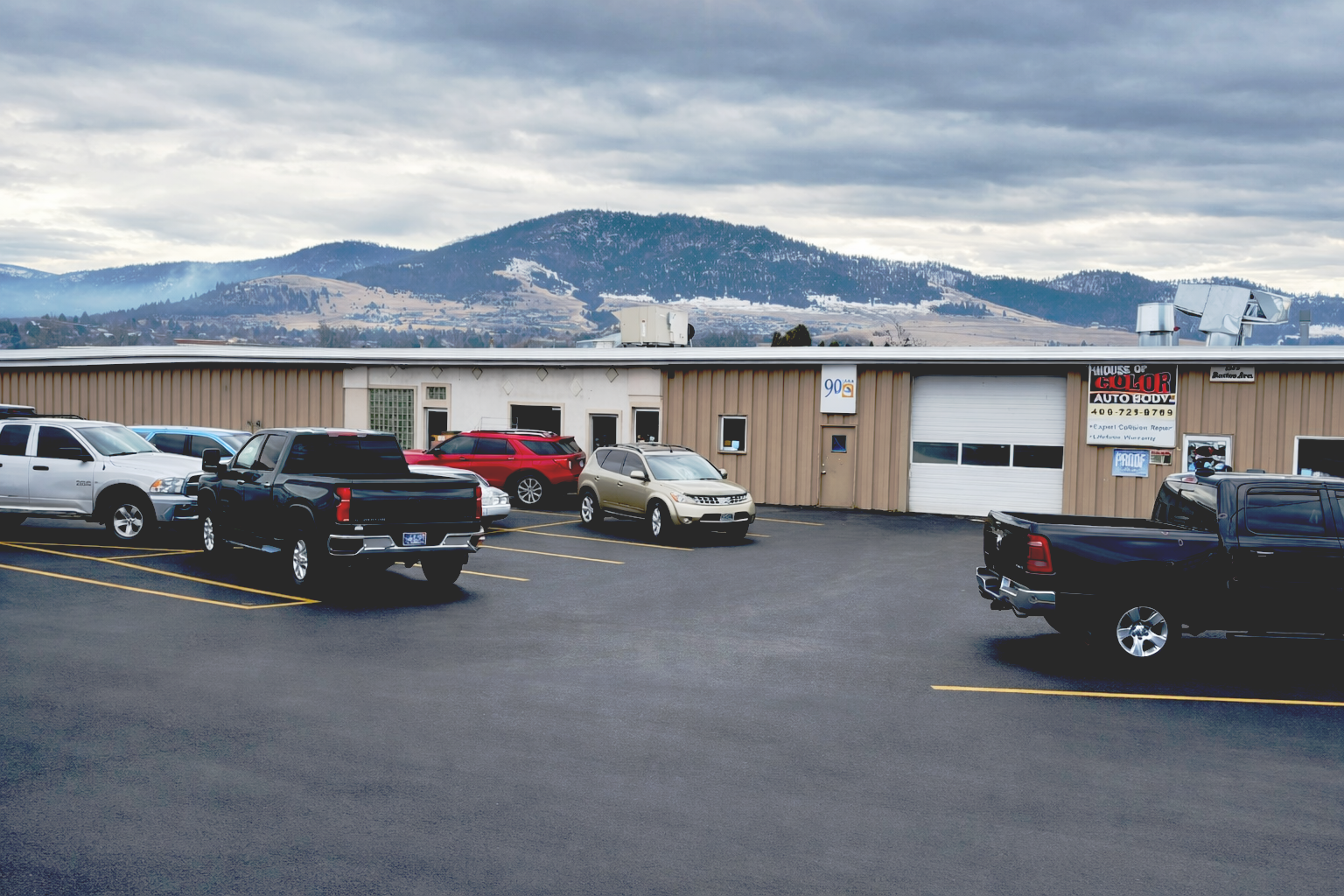 Cars parked in front of a tan building, House of Color Auto Body, mountains in the background under a cloudy sky.