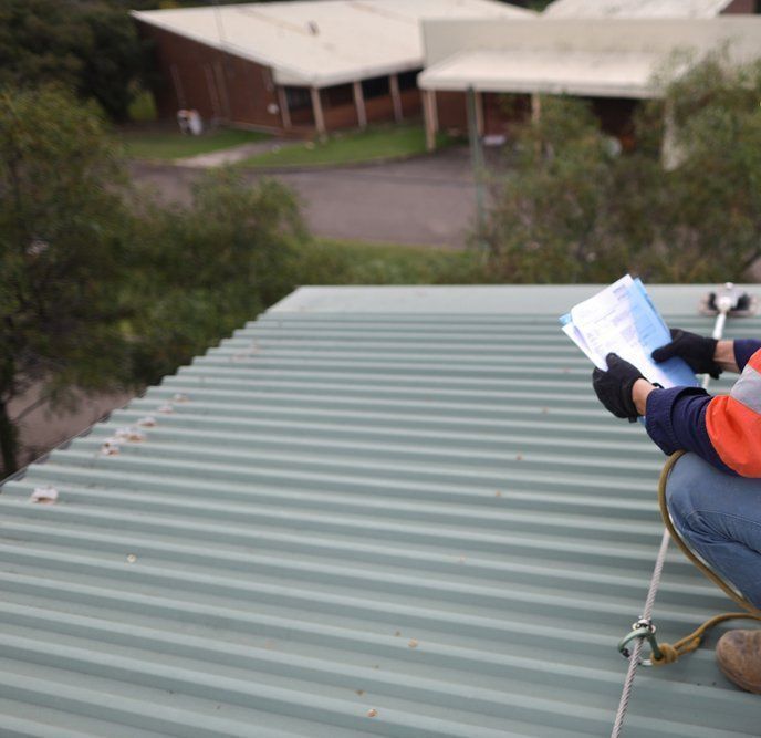 A Man is Kneeling on a Roof Looking at a Piece of Paper — Goulburn Roofing In Goulburn, NSW
