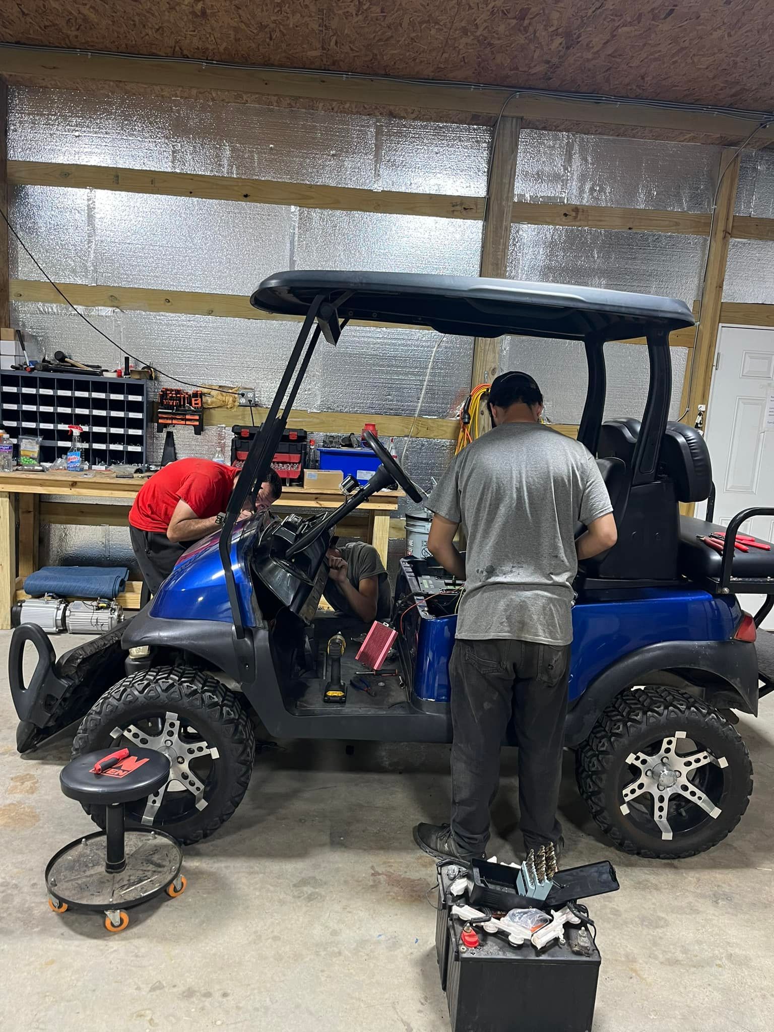 Two people working on a blue golf cart in a garage. One person is standing near the cart, and the other is bent over it.