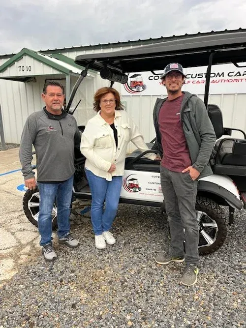 Three people stand by a white golf cart in front of a business with a sign.