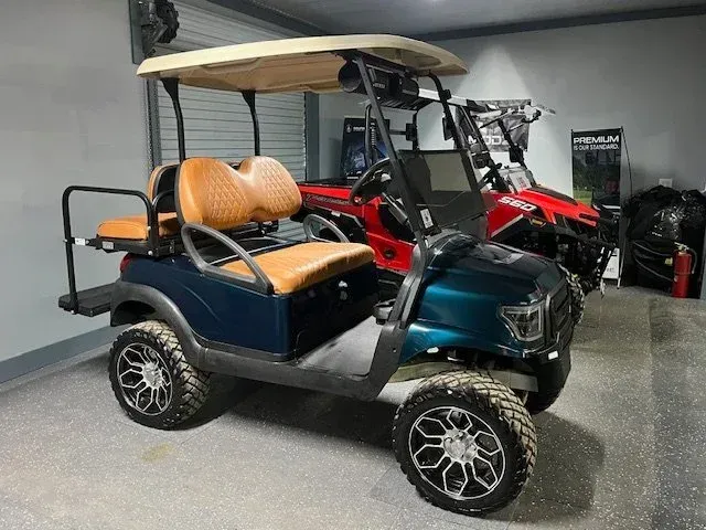 Dark blue golf cart with tan seats and a beige roof. Black wheels and a red vehicle in the background.