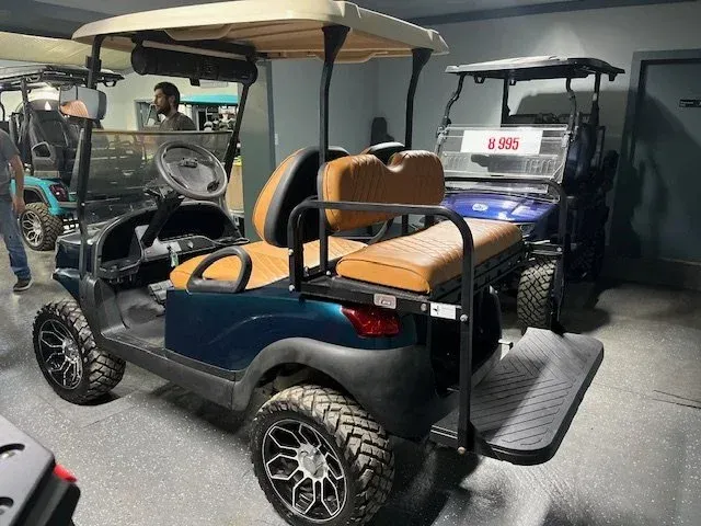 Golf cart, teal body, brown seats, black wheels. Other carts and people in background.