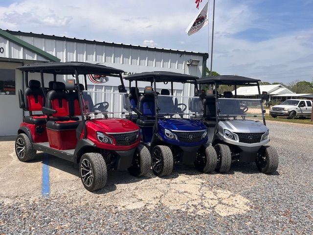 Three modern golf carts in red, blue, and silver parked in a row outside a white building under a clear blue sky.