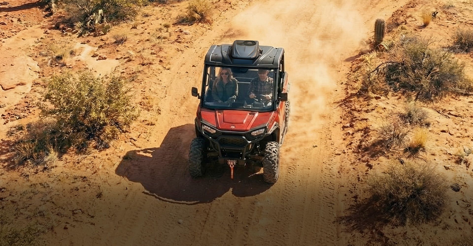 Red off-road vehicle driving on a dusty dirt trail through dry, sparse brush.