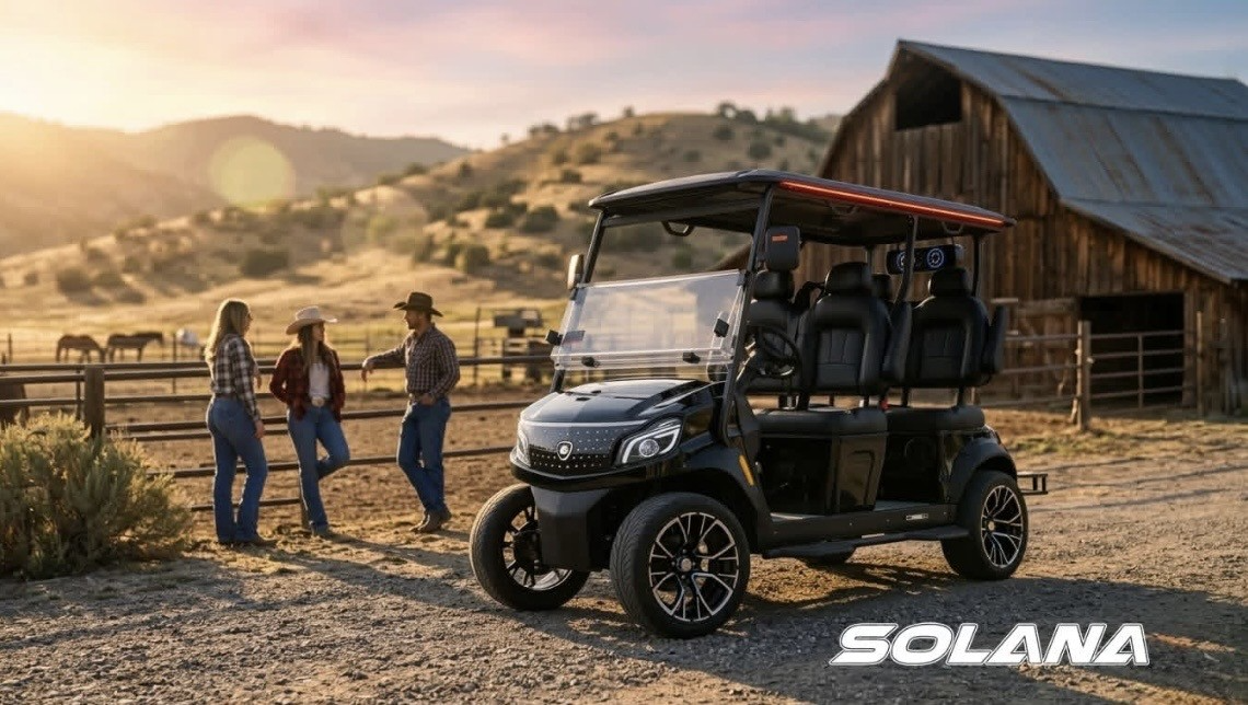 A black Solana electric cart parked on a dirt path by a wooden barn, with three people standing and talking nearby.