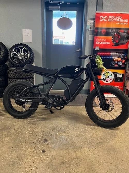 Black electric motorbike indoors. Thick tires, black frame, in front of a door and tires.
