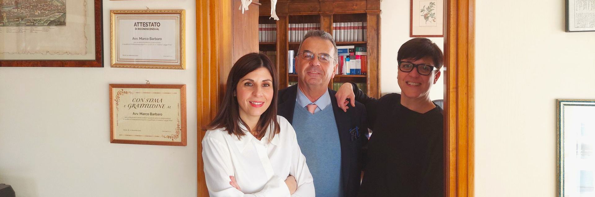 Tre persone posano sulla soglia di una porta: due donne e un uomo, sorridenti. Certificati incorniciati sono appesi al muro.