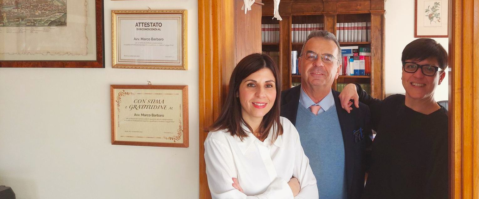 Tre persone sono in piedi sulla soglia di una porta: una donna, un uomo e una persona con gli occhiali. Sullo sfondo una libreria, documenti incorniciati sulla parete.