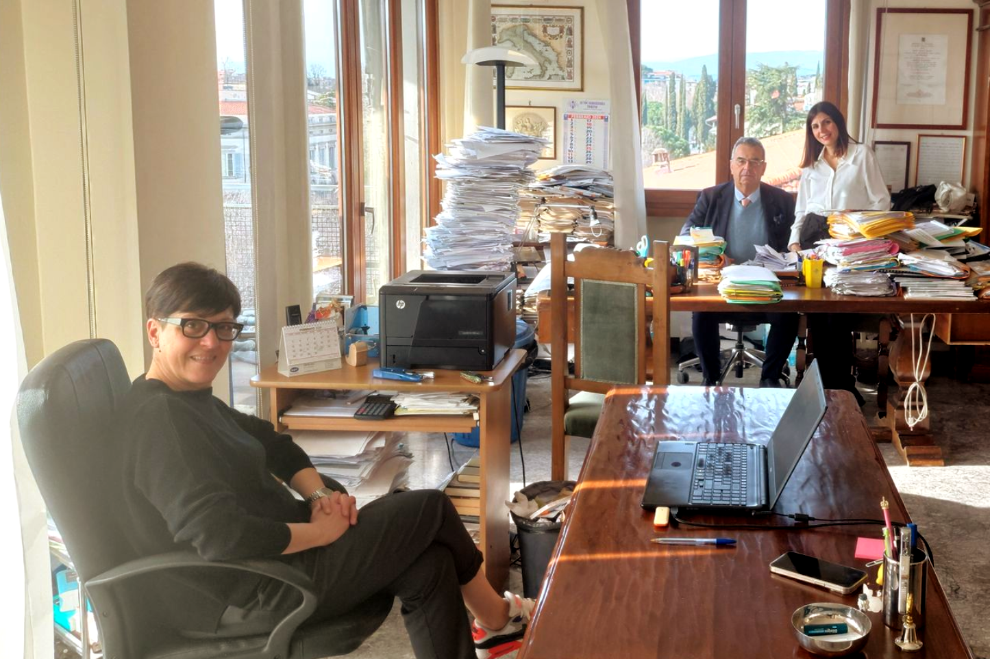 Una donna è seduta su una sedia da ufficio, sorridente. Altre due persone sono sedute alla scrivania ingombro di stampanti, documenti e una finestra con vista.