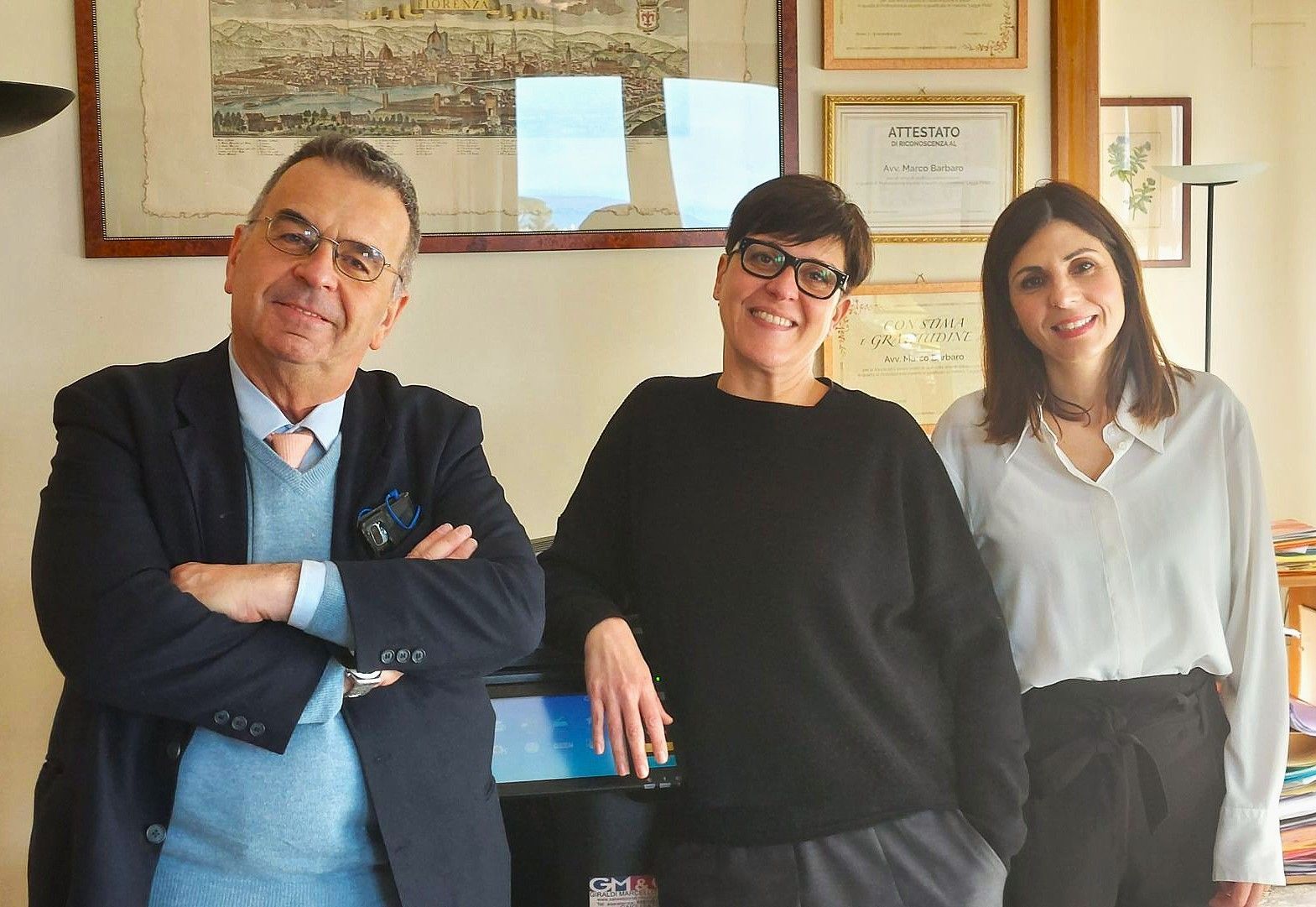 Tre persone in posa insieme in un ufficio. Un uomo con le braccia incrociate, due donne, tutti sorridenti. Opere d'arte e diplomi appesi al muro.