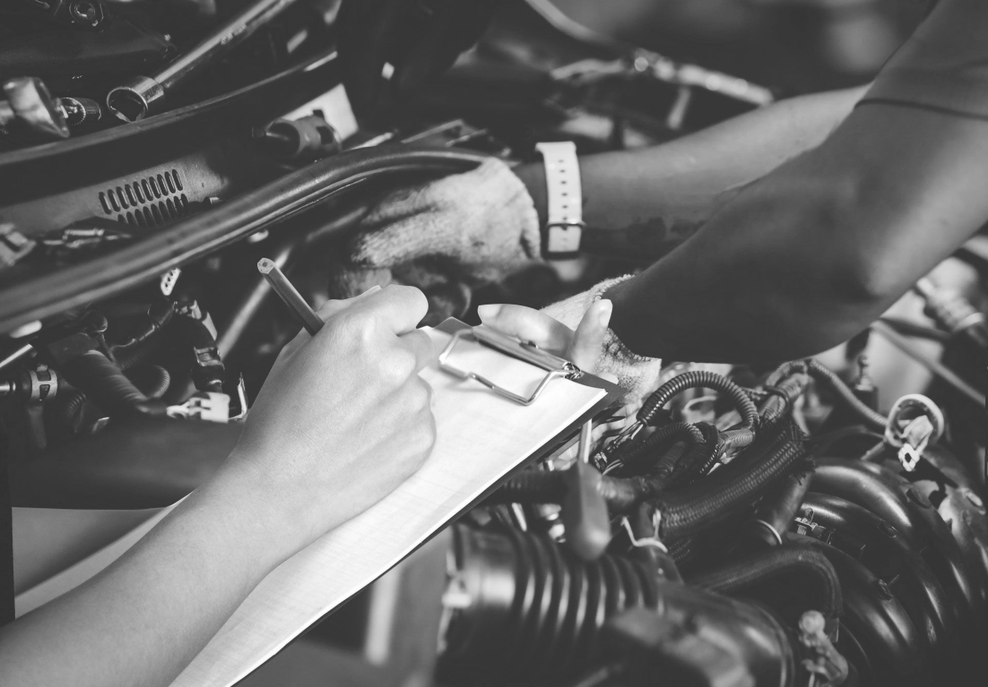 A person wearing a glove works on a car engine while another person takes notes on a clipboard.