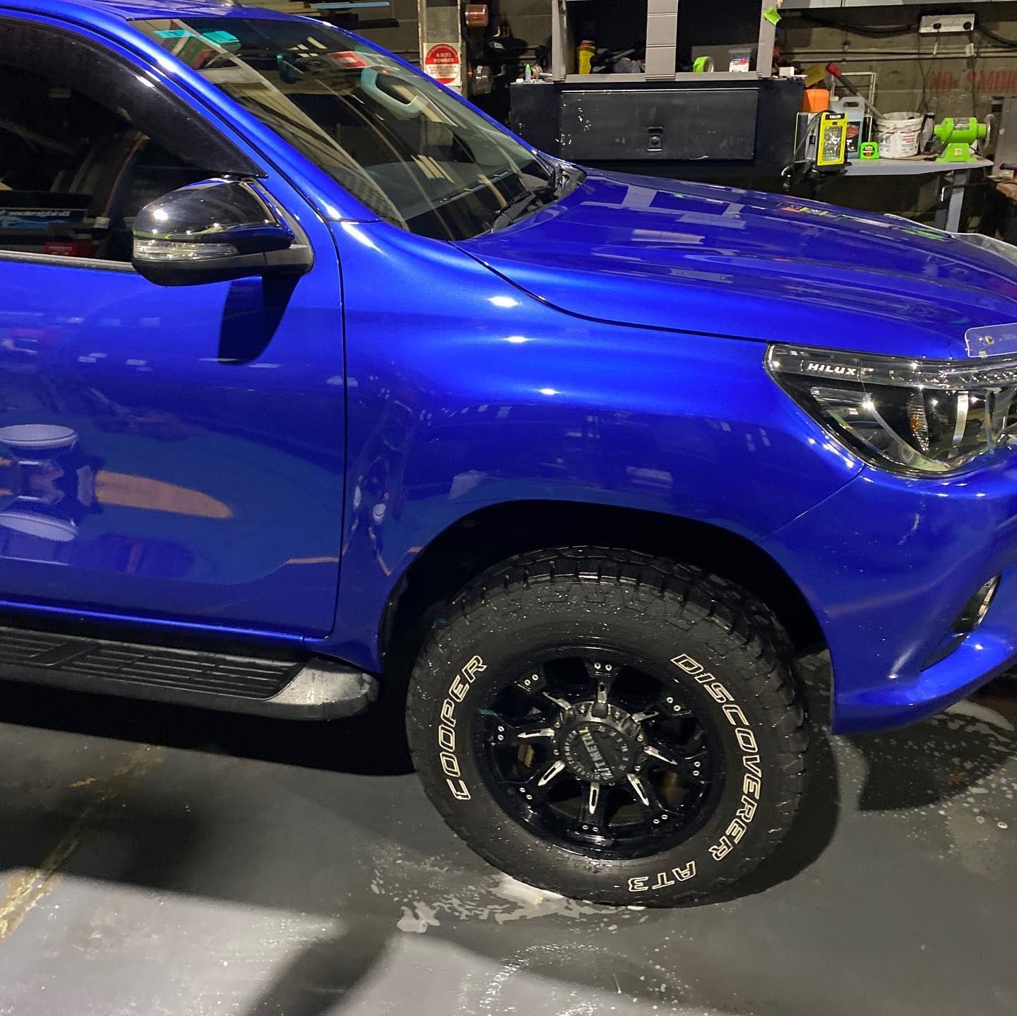 A bright blue pickup truck is parked indoors, showcasing its front wheel with a black rim and textured all-terrain tire.