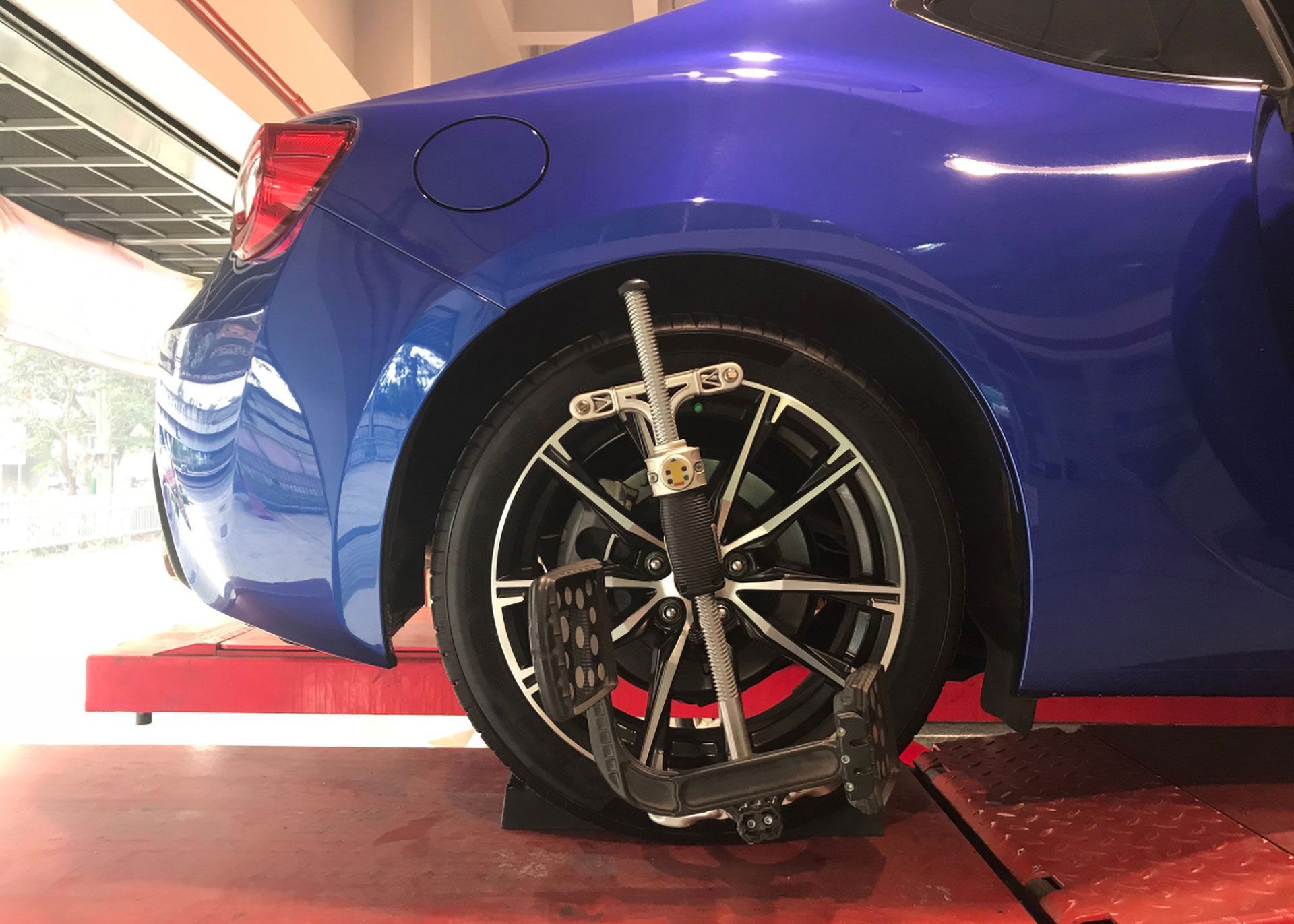 A blue car parked on a red alignment lift with a wheel alignment sensor attached to the rear tire.
