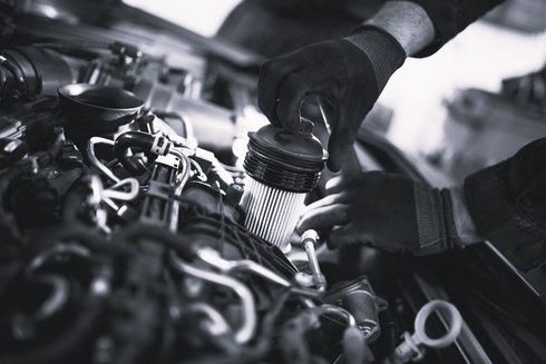 A mechanic in black gloves performs maintenance on a car engine, holding a cylindrical oil filter element.