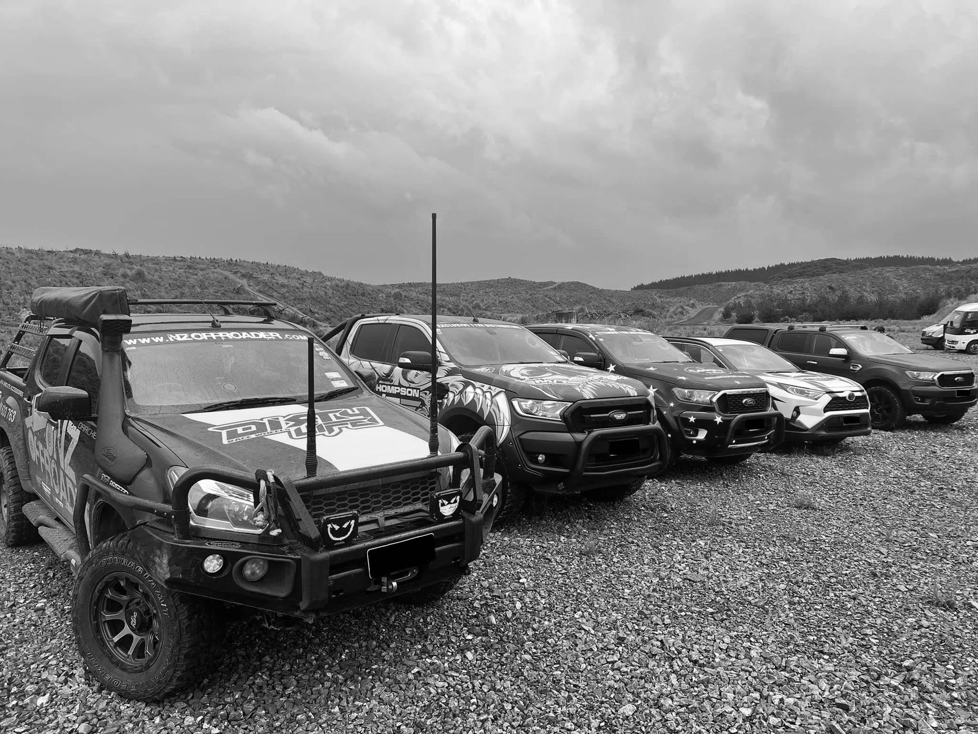 A line of lifted pickup trucks parked on a gravel surface under a cloudy sky.