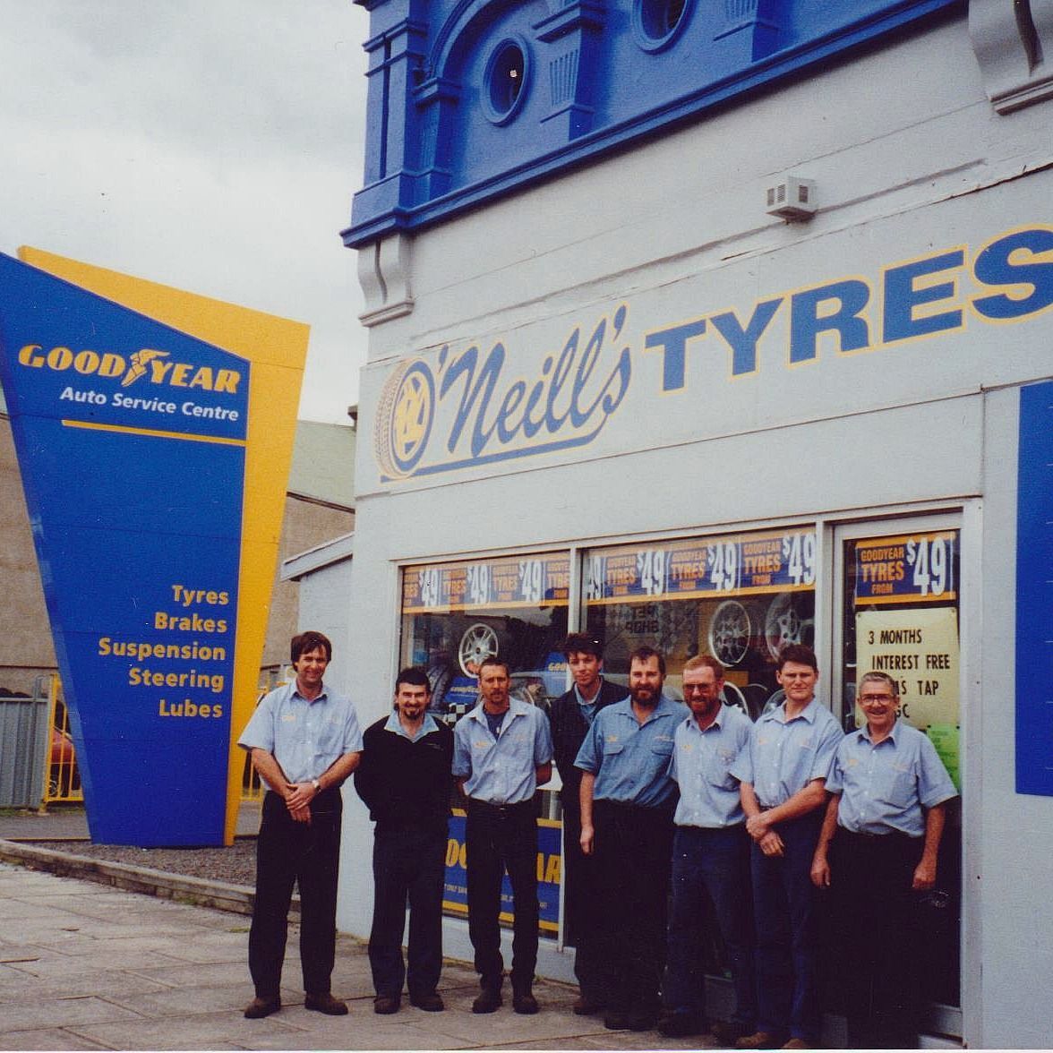 A group of eight workers in blue shirts standing in front of O'Neill's Tyres, a Goodyear auto service center.