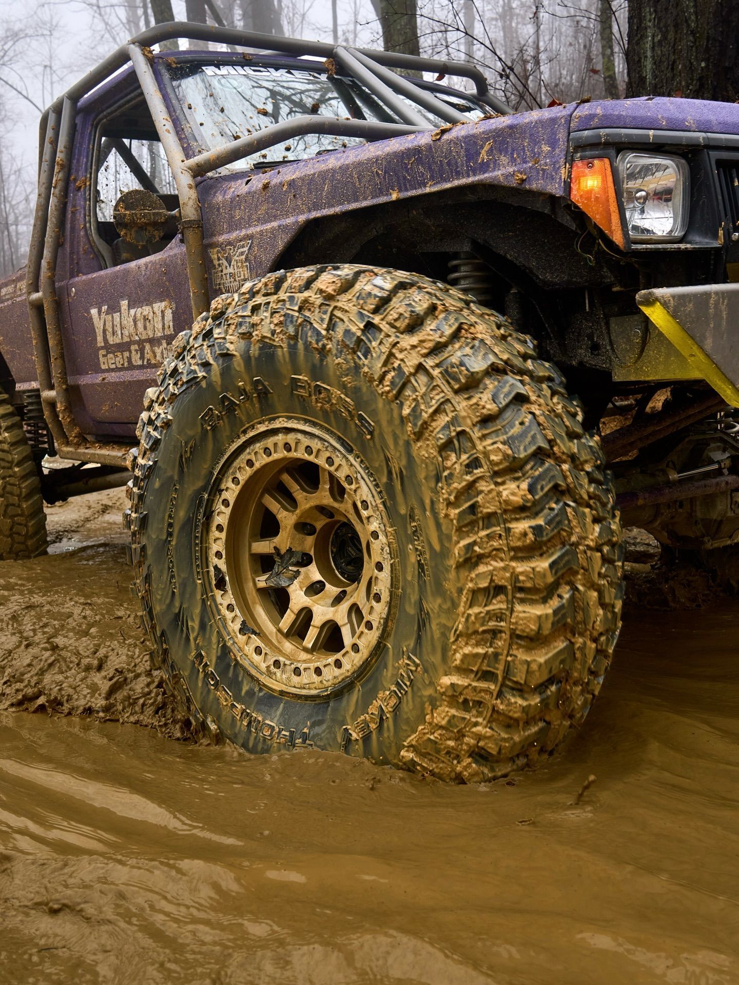 A purple off-road vehicle with large, mud-caked tires drives through deep mud in a forest.