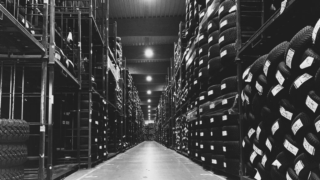 A monochrome shot of a long warehouse aisle lined with floor-to-ceiling metal shelving stacked with many car tires.